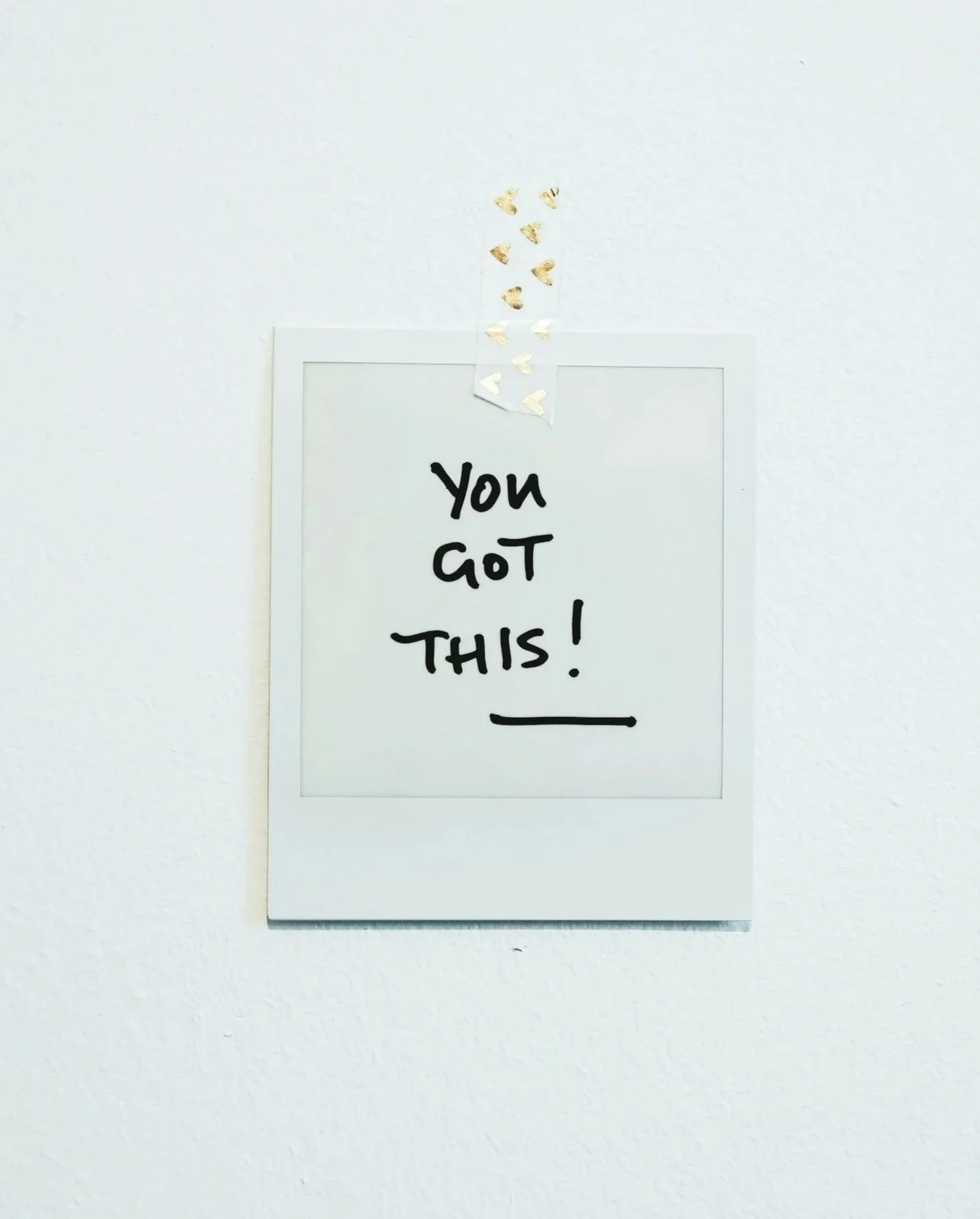 A whiteboard with the phrase 'You Got This!' written in black marker, decorated with gold mini heart-shaped accents taped above.