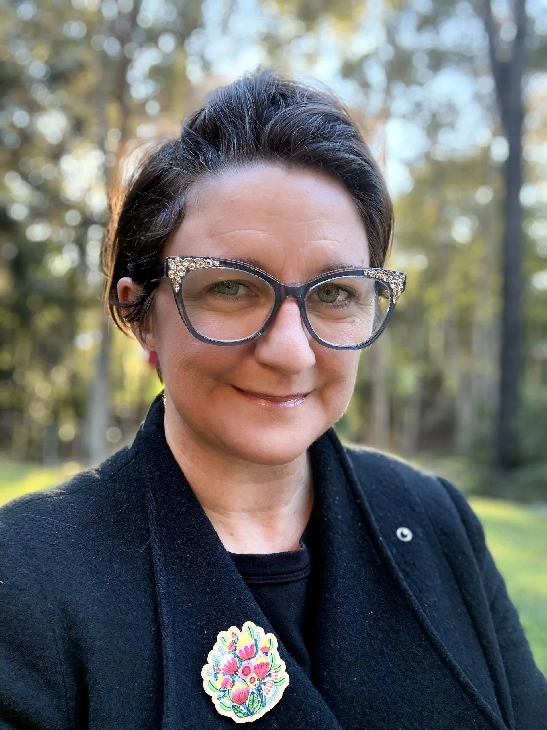 A woman with short dark hair, wearing glasses with decorative rhinestones on the frames, smiling outdoors with a blurred forest background, wearing a black outfit and a decorative floral sticker on her chest.
