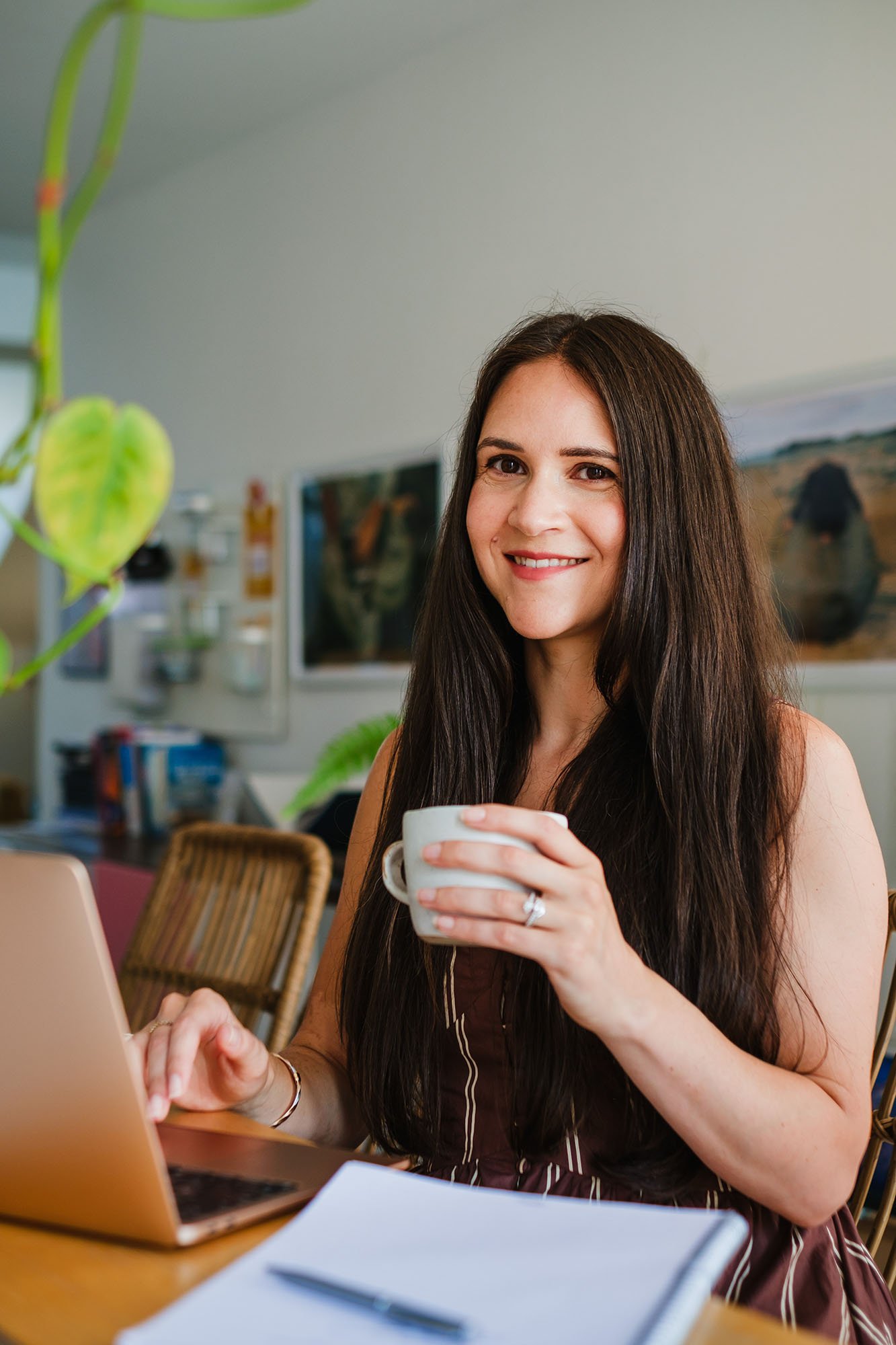 Woman with long dark hair smiling while holding a white coffee cup, sitting indoors with a laptop and notebook in front of her.