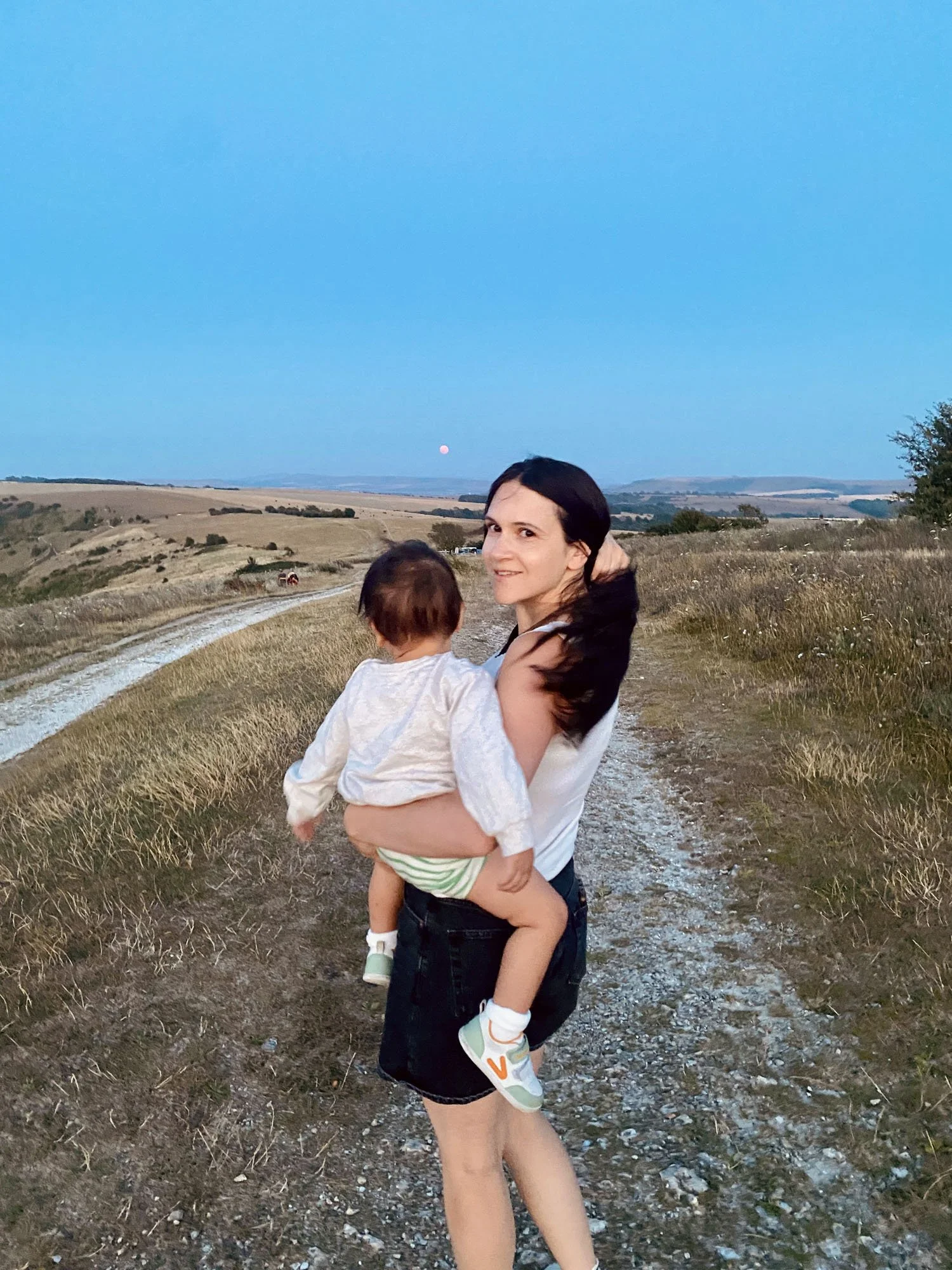A woman with dark hair carrying a young child on a dirt trail in a rural landscape during dusk, with a moon visible in a clear sky and rolling hills in the background.