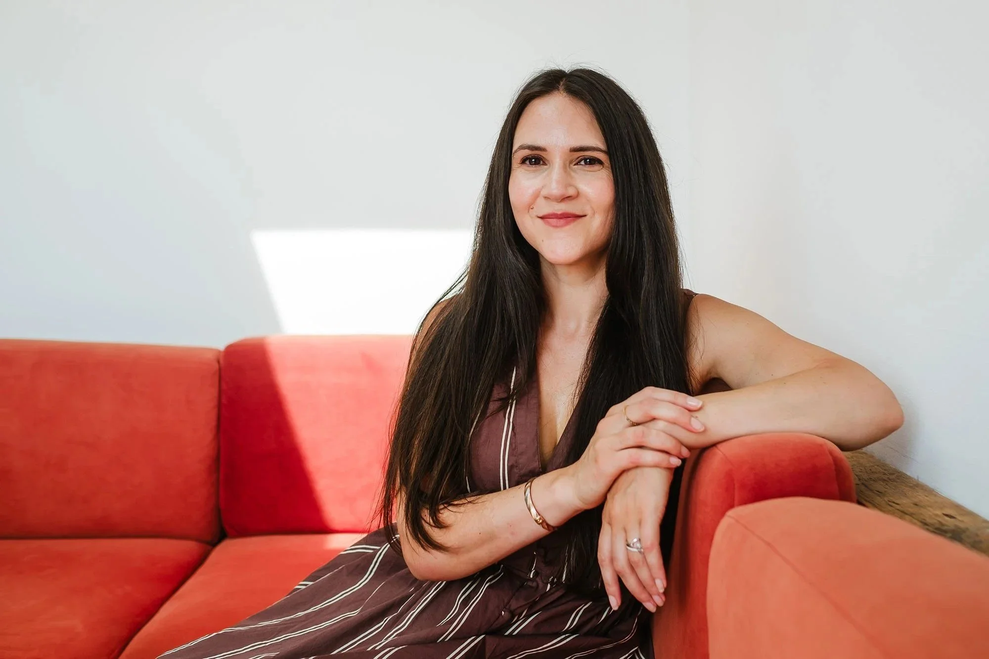 A woman with long dark hair and a slight smile sitting on a red sofa, with a white wall in the background.