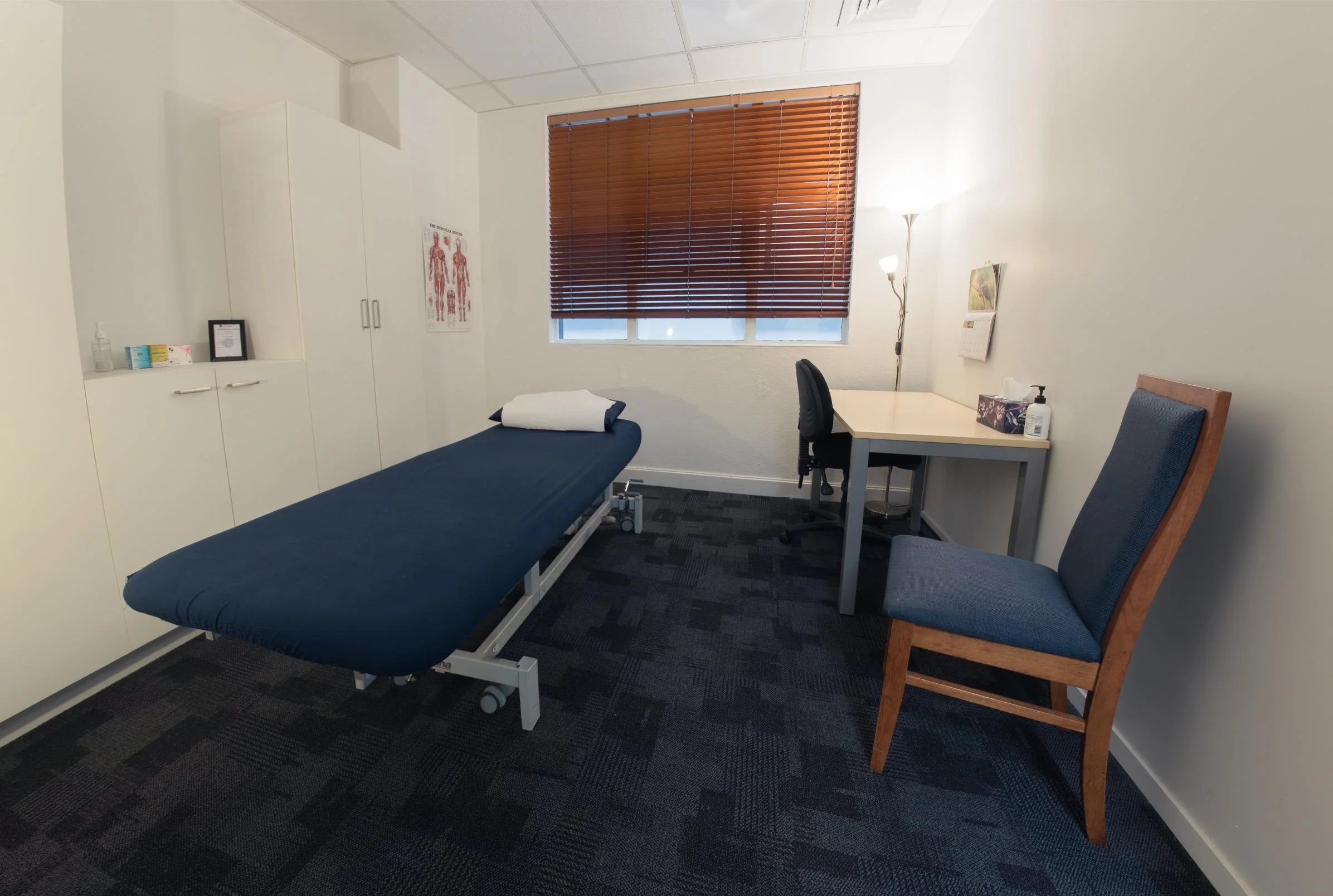 Medical exam room with an examination bed, a desk with a chair, a bookshelf, a lamp, and medical supplies on a cabinet. The room has a window with brown blinds.