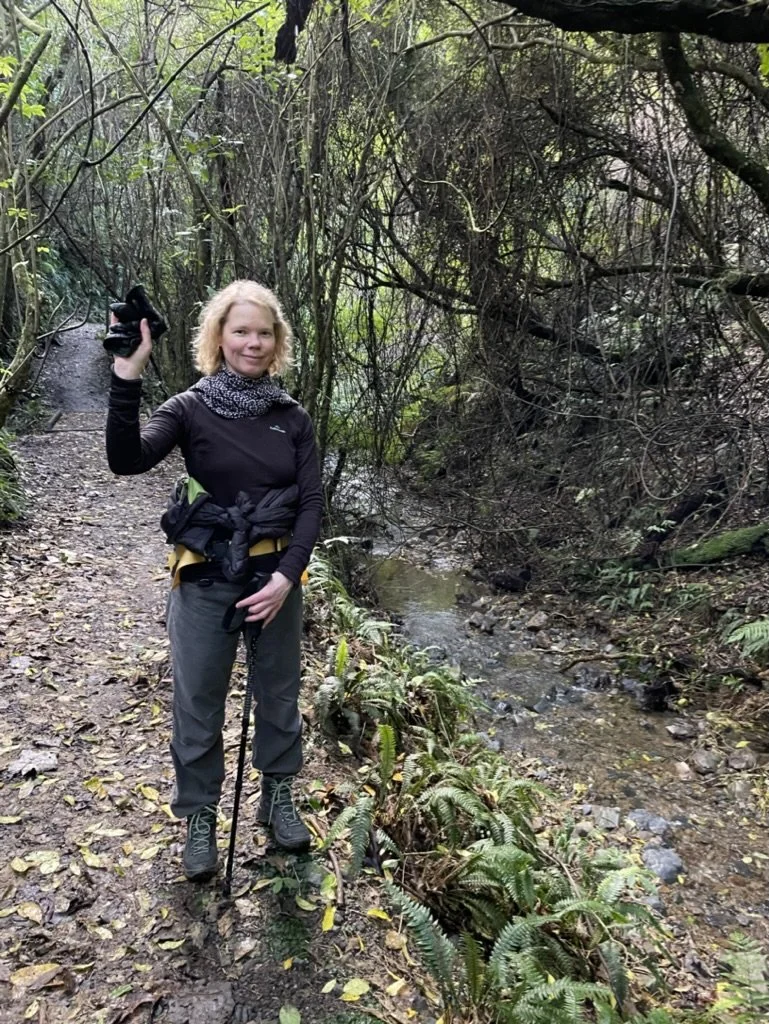 A woman with blonde hair standing on a trail in a forest, holding a camera in her right hand and a walking stick in her left. She is dressed in outdoor hiking gear surrounded by dense trees and foliage, next to a small creek.
