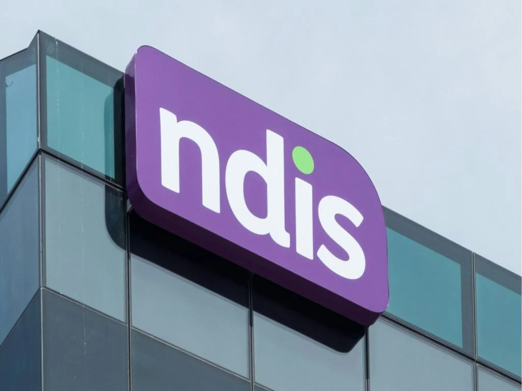A corporate building with a purple sign displaying the word "ndis" in white lowercase letters, with a small green dot above the letter "i".