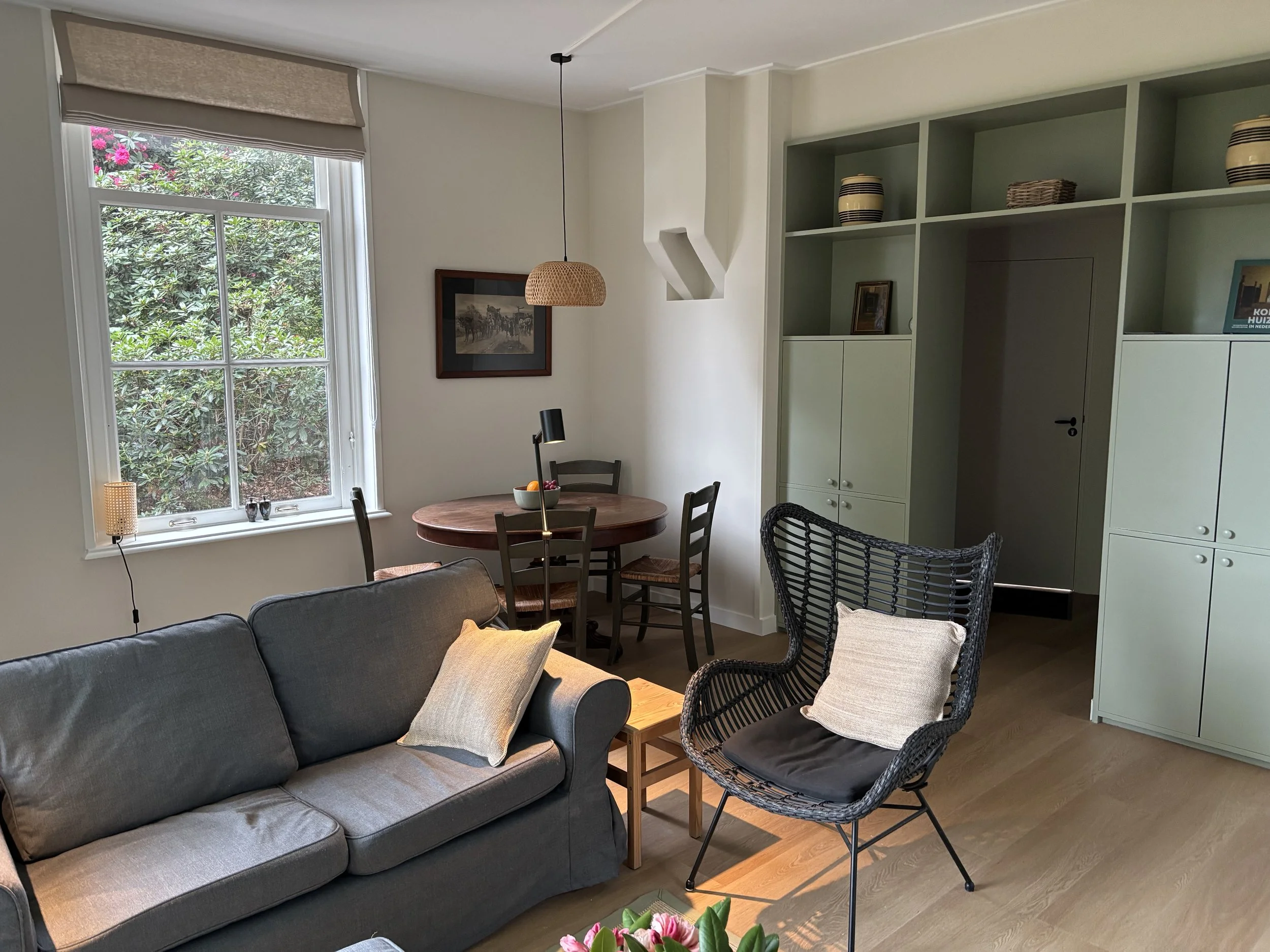 Woonkamer met grote venster, bank, ronde eettafel met stoelen, wandkast met decoraties, middelhoge museumwerk en zitkussen, houten vloer.