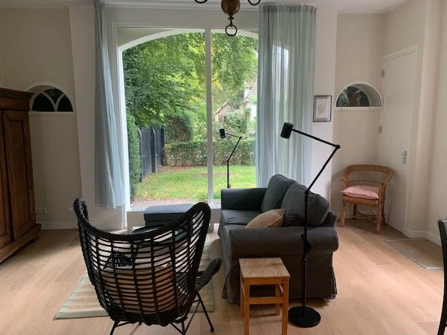 Gezellig woonkamer met grote ramen, groene tuin buiten, zithoek met bank, houten bijzettafel, leeslampen en een rieten stoel.