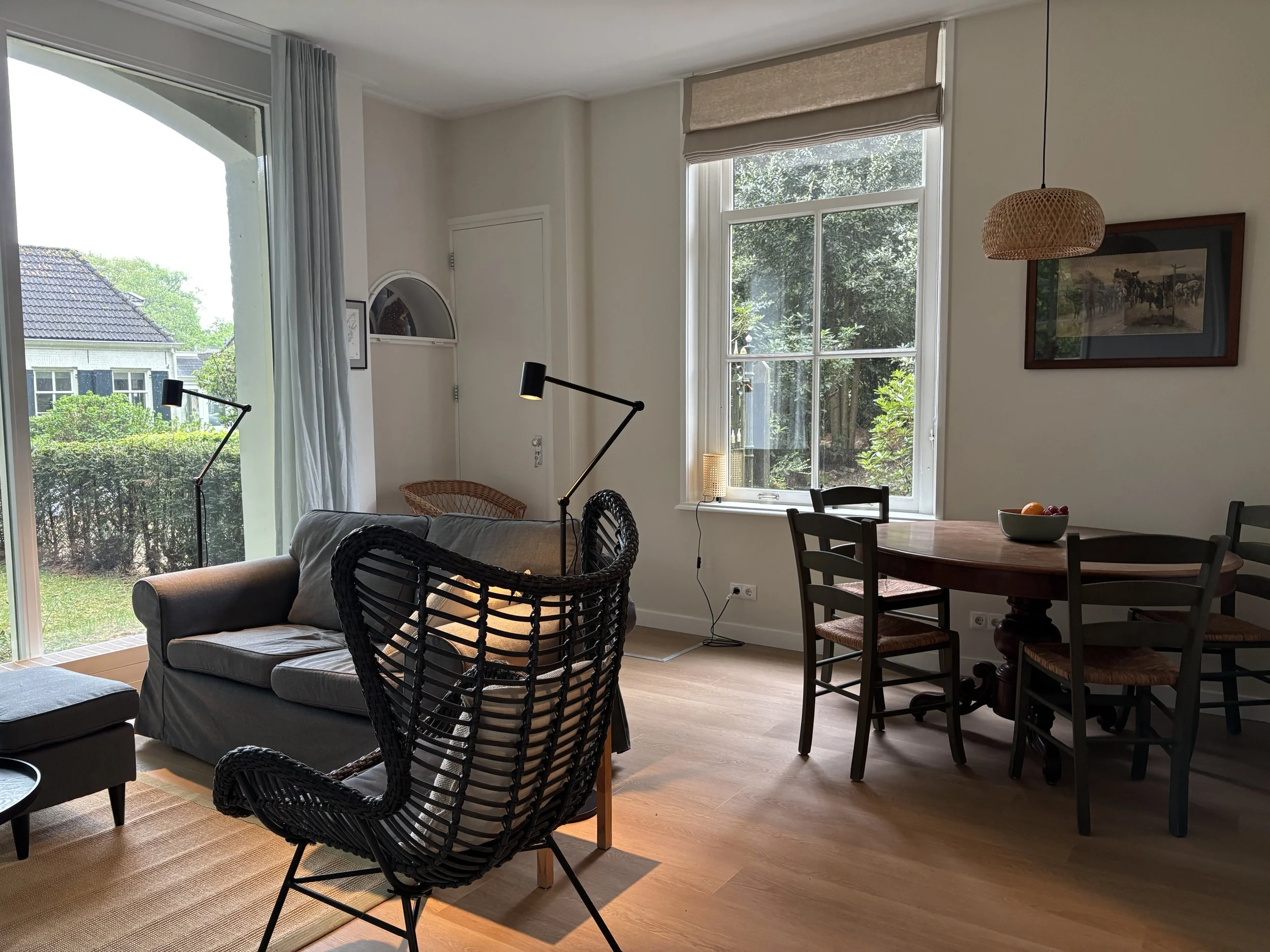 Interieur van een woonkamer met grote ramen, een bank, een eettafel, en diverse lampen, met uitzicht op de tuin buiten.