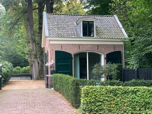 Kleine huisje met dakraampje, groene luiken en een oprijlaan, omgeven door bomen en heggen.