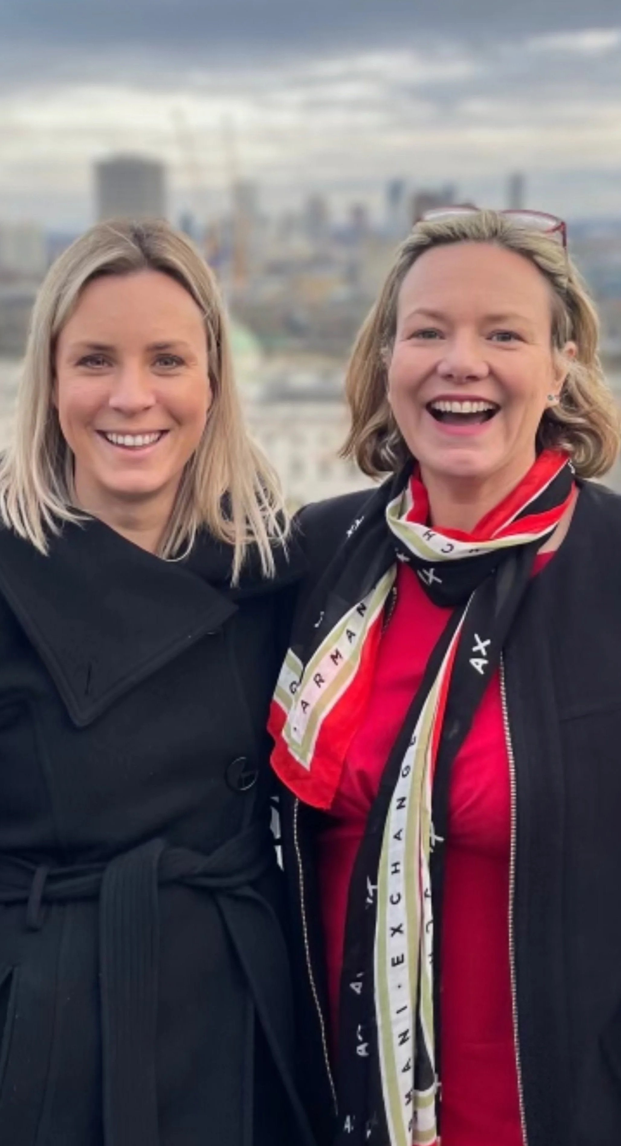 Two smiling women standing outdoors with a city skyline in the background on a cloudy day.