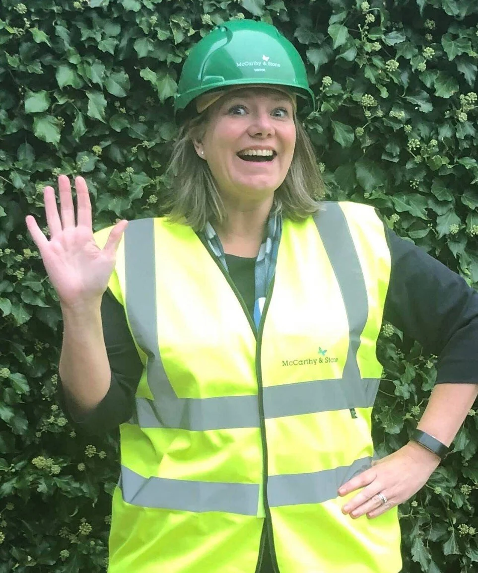 A woman wearing a green safety helmet and a yellow reflective safety vest, waving with her right hand, standing in front of a leafy green wall.