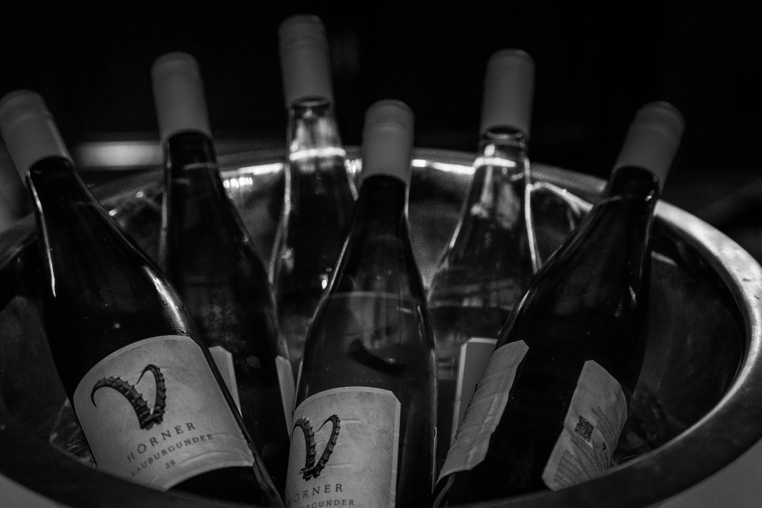 Schwarz-weiß-Foto, Weinflaschen im Weinregal, einige Flaschen zeigen Details auf den Etiketten, dunkles Hintergrund.