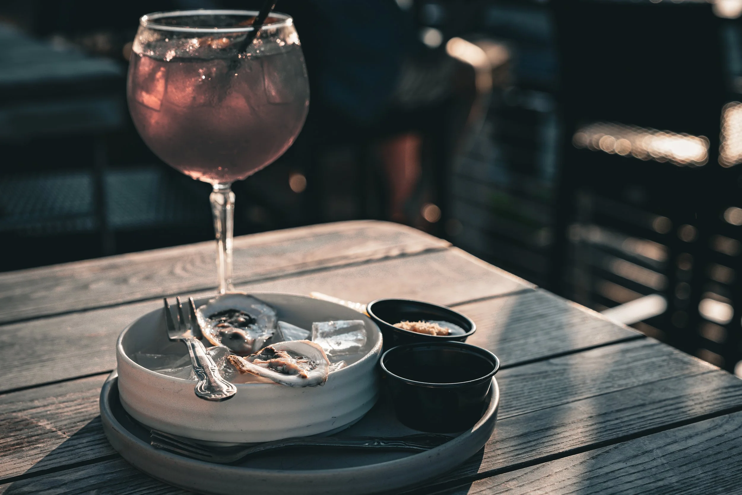 Ein Gartentisch mit einer Schüssel Austern, zwei kleine Schälchen mit Soße und ein Glas Roséwein mit Eiswürfeln und einem Strohhalm, im Hintergrund ein dunkler, unscharfer Gartenbereich bei Sonnenuntergang.