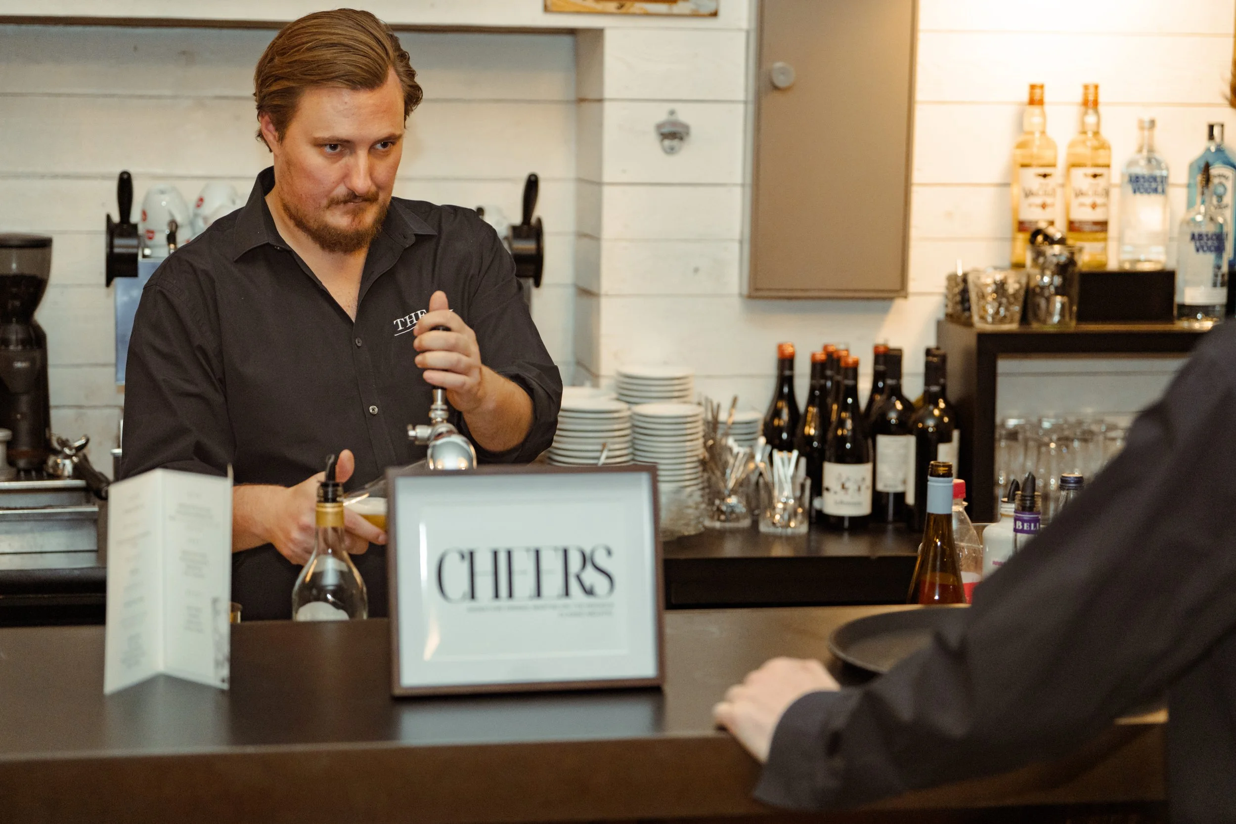 Ein Barkeeper bereitet Getränke hinter der Theke zu, im Vordergrund eine Person, die an der Bar sitzt.