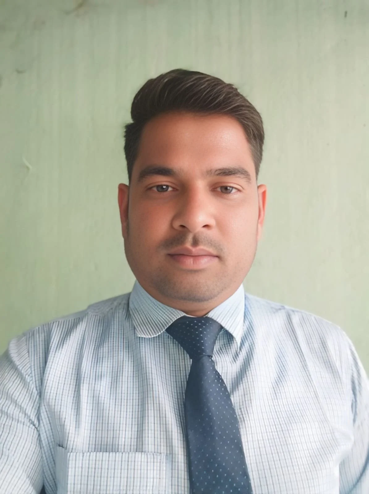 A young man with short dark hair, wearing a light-striped dress shirt and a navy blue tie with small dots, standing in front of a plain green wall.
