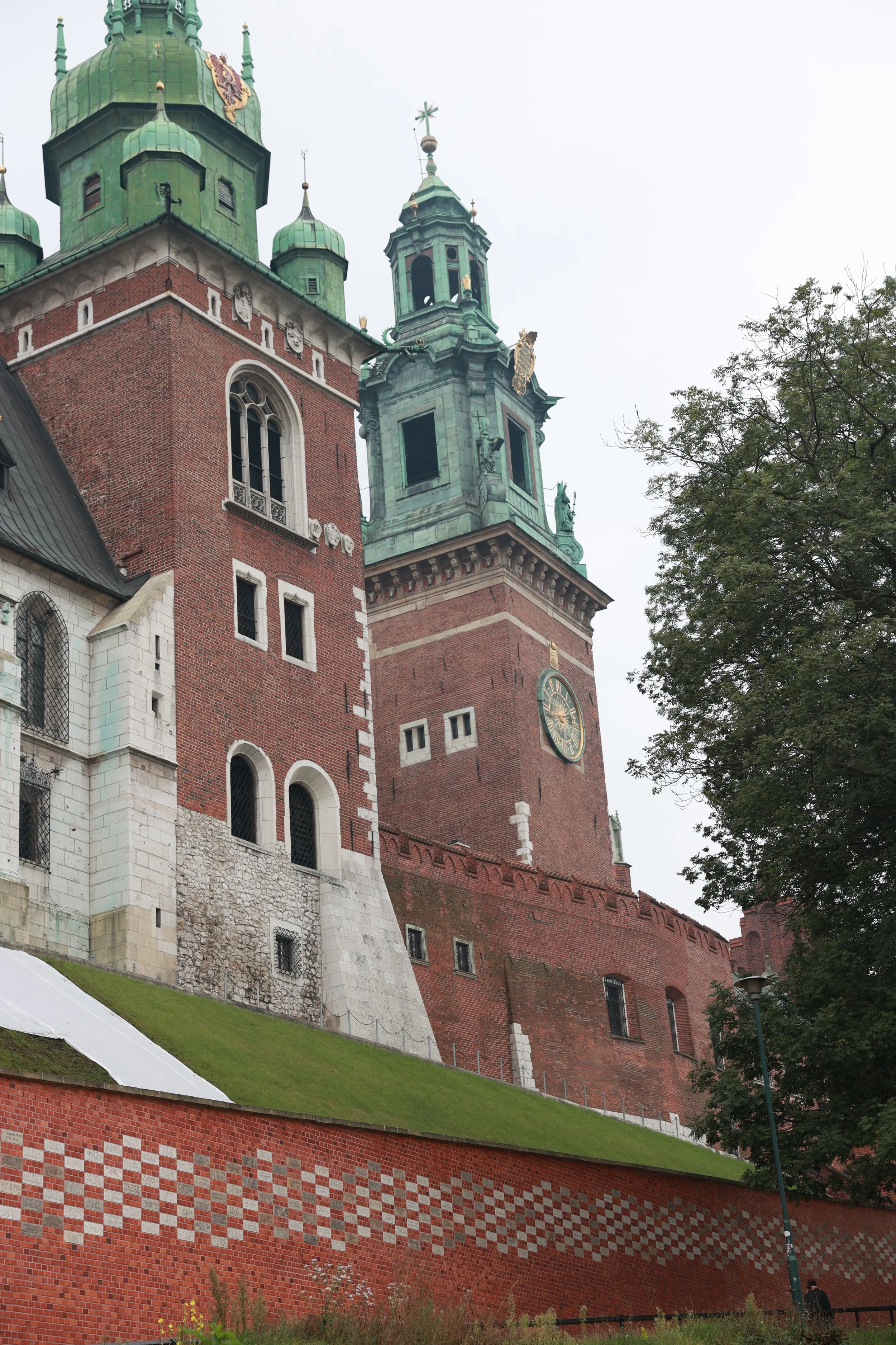 A large historic building with red brick walls, green domed towers, a clock, and decorative statues, surrounded by trees and a grassy hill with a patterned brick wall at the bottom.