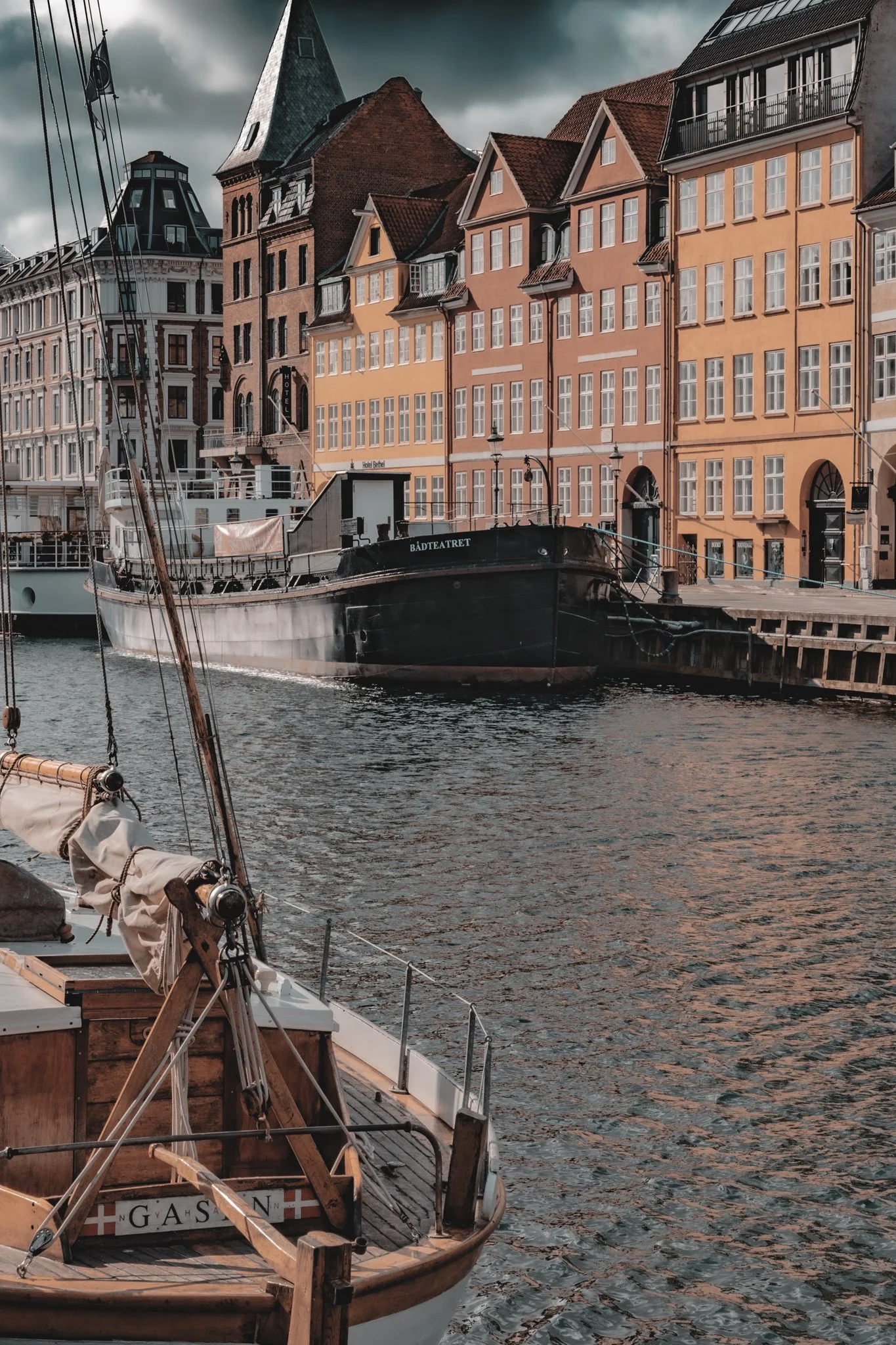 A view of colorful European-style buildings beside a marina with boats, including a sailboat in the foreground and a larger boat named BADTEATRET docked nearby. The buildings have multiple stories with large windows and steep roofs, some with small balconies. The sky is cloudy.