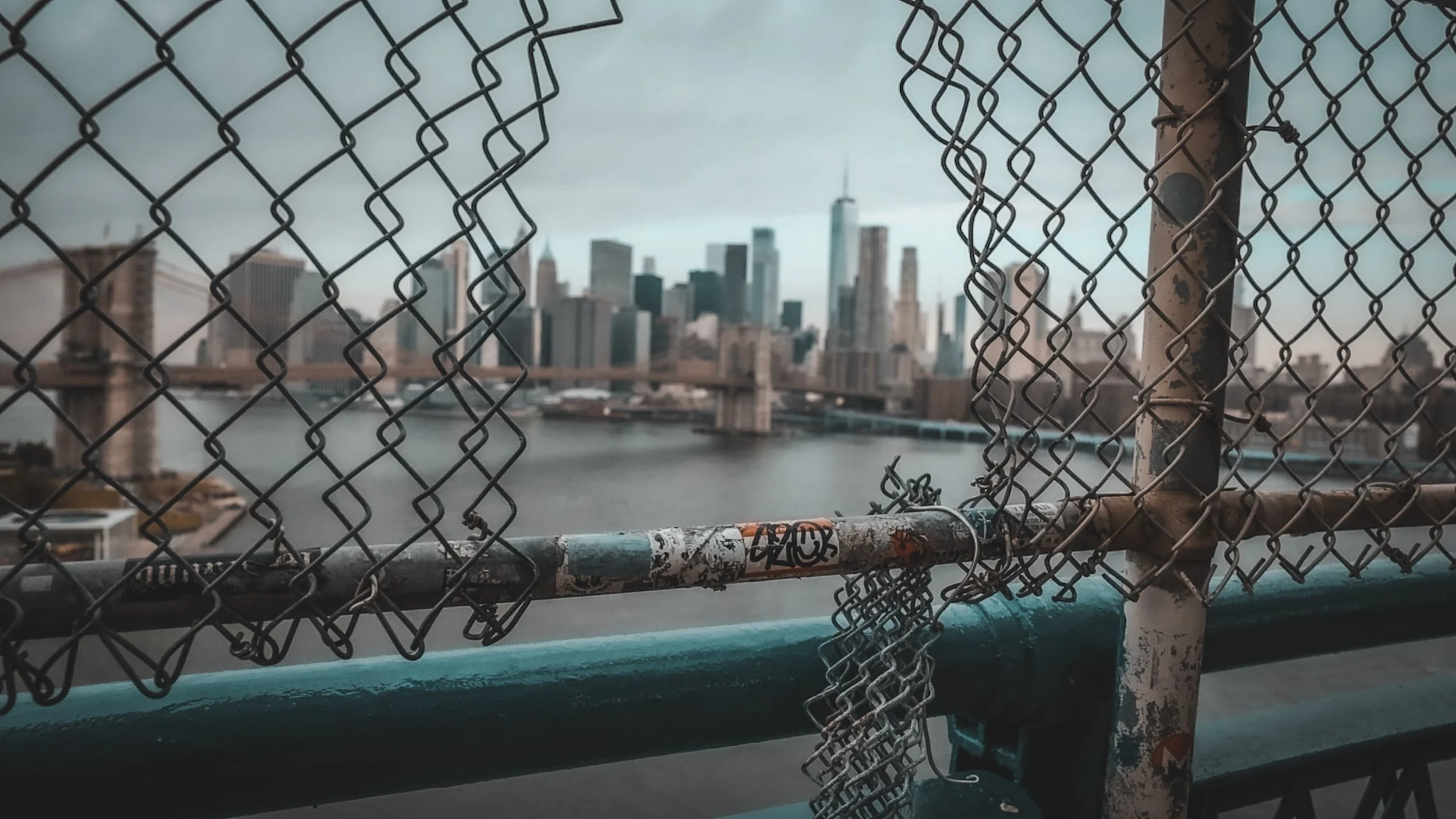 City skyline viewed through a chain-link fence on a bridge, with overcast sky.