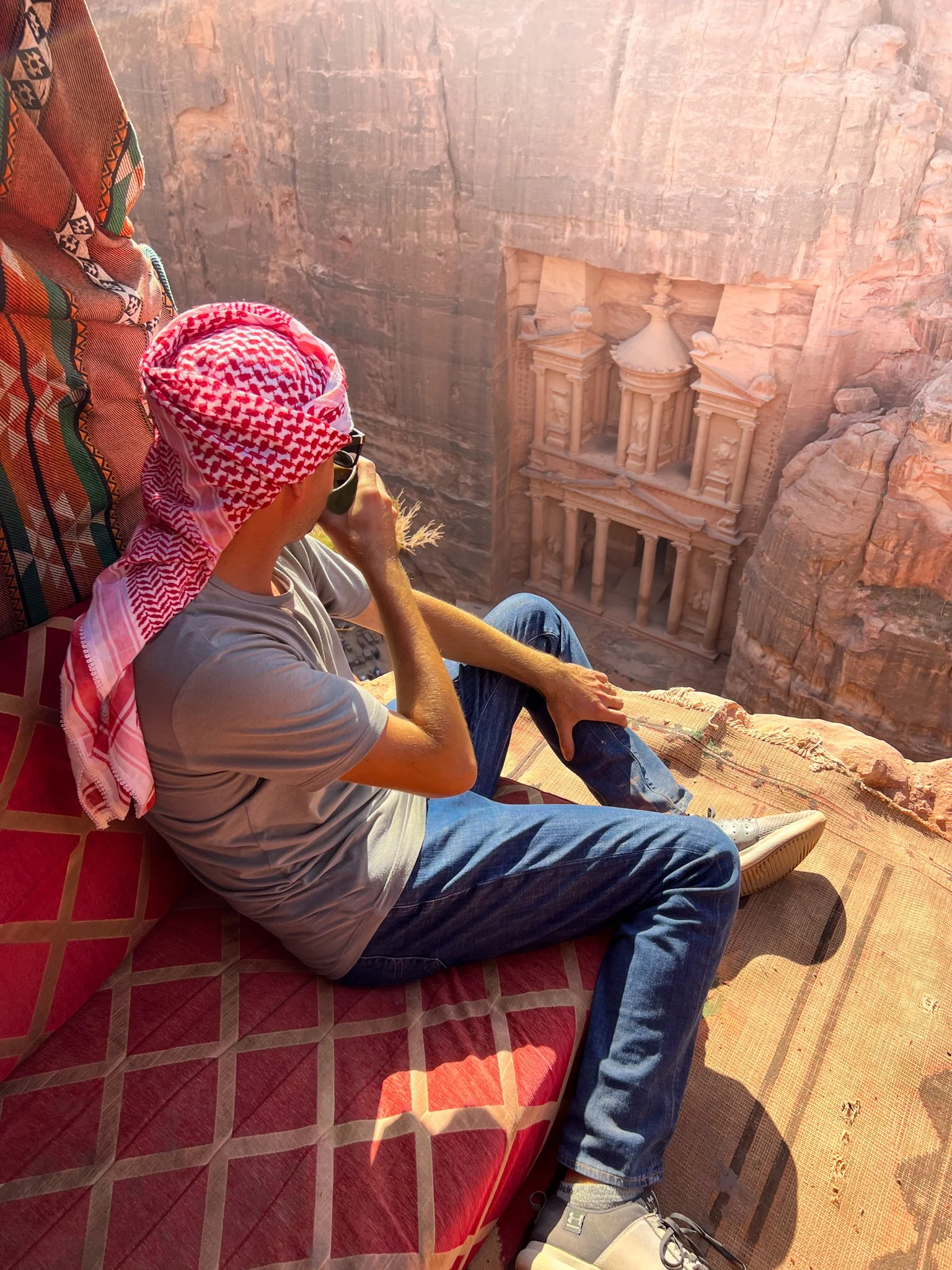 A man sitting on a patterned red cushion on the floor, looking through binoculars at the ancient rock-carved city of Petra, Jordan, which is partially visible in the canyon wall.