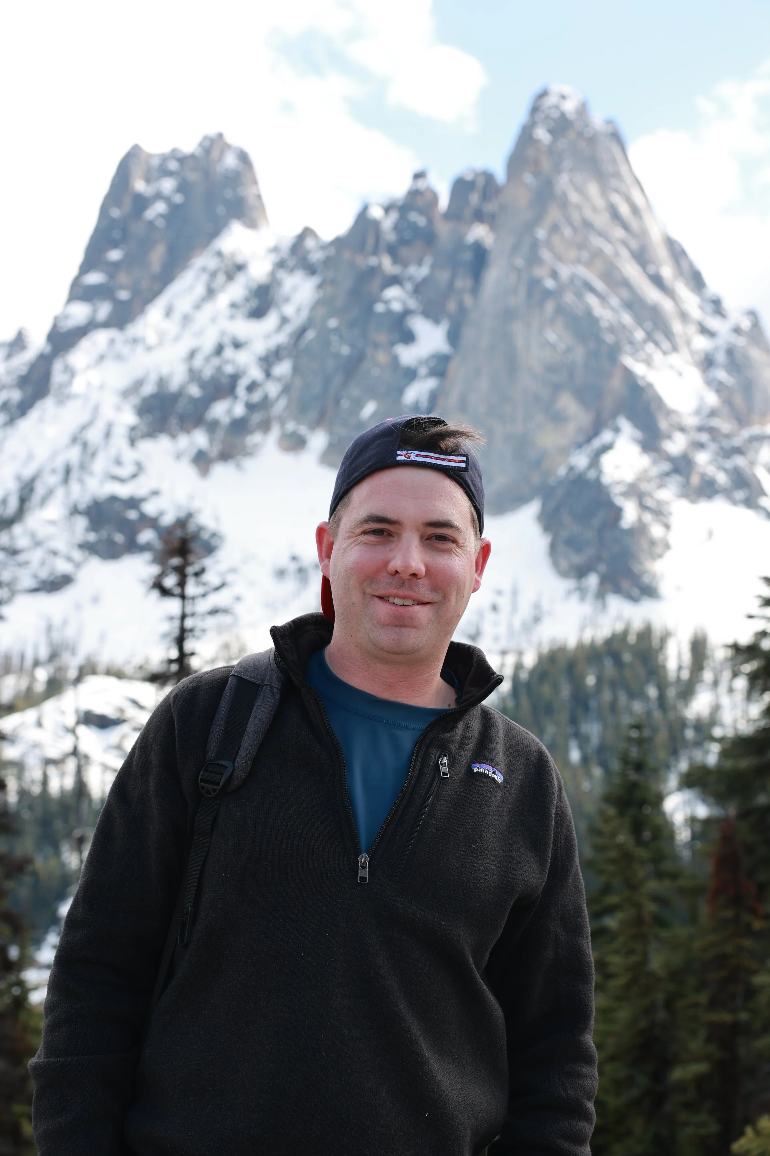 A smiling man wearing a black jacket and a baseball cap, standing outdoors with snow-covered mountains and trees in the background.
