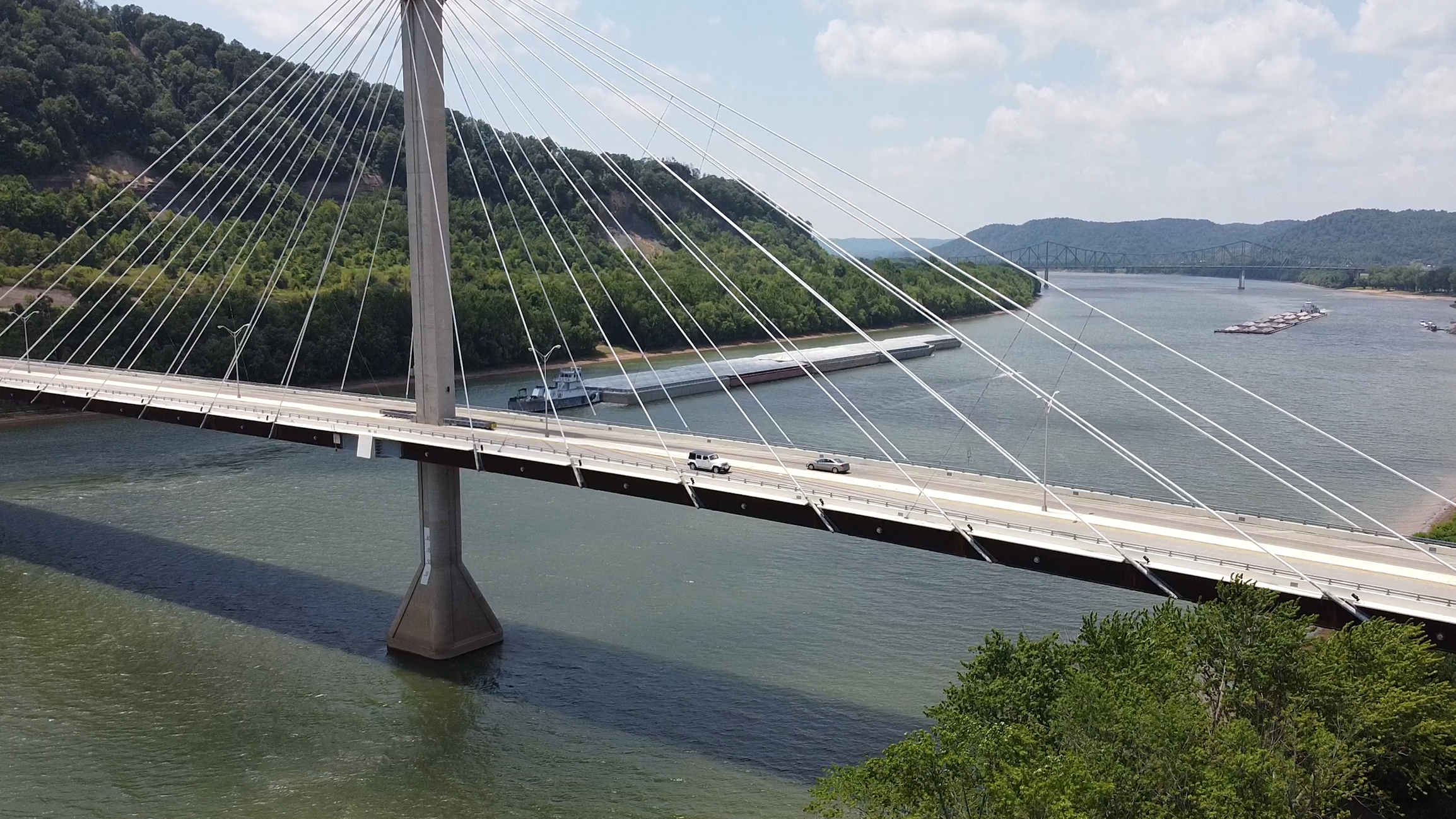 A modern cable-stayed bridge spanning a wide river, with a green hillside in the background, and a small boat near the bridge.