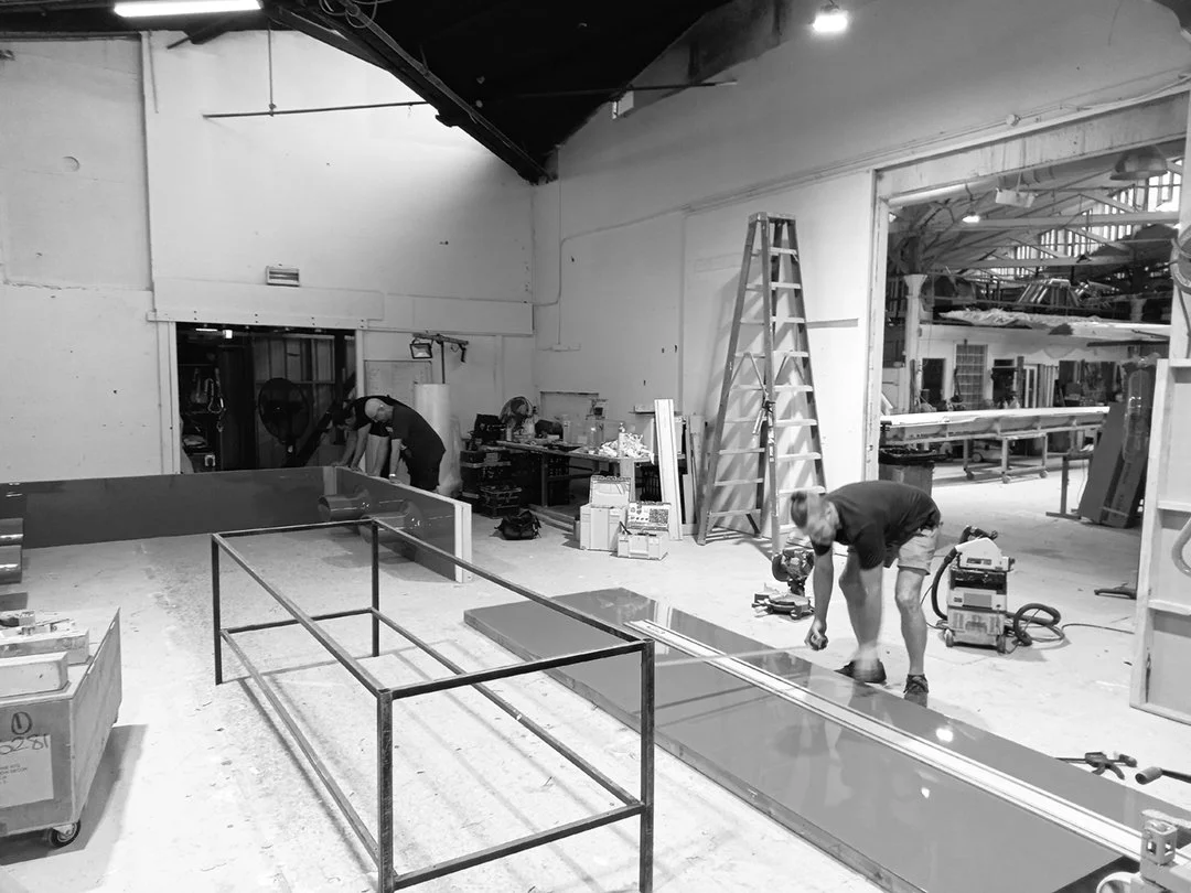 Workers assembling or repairing a large structure in a workshop with tools, a ladder, and various equipment around. HR Productions workshop space. Hire Custom built, props, sydney