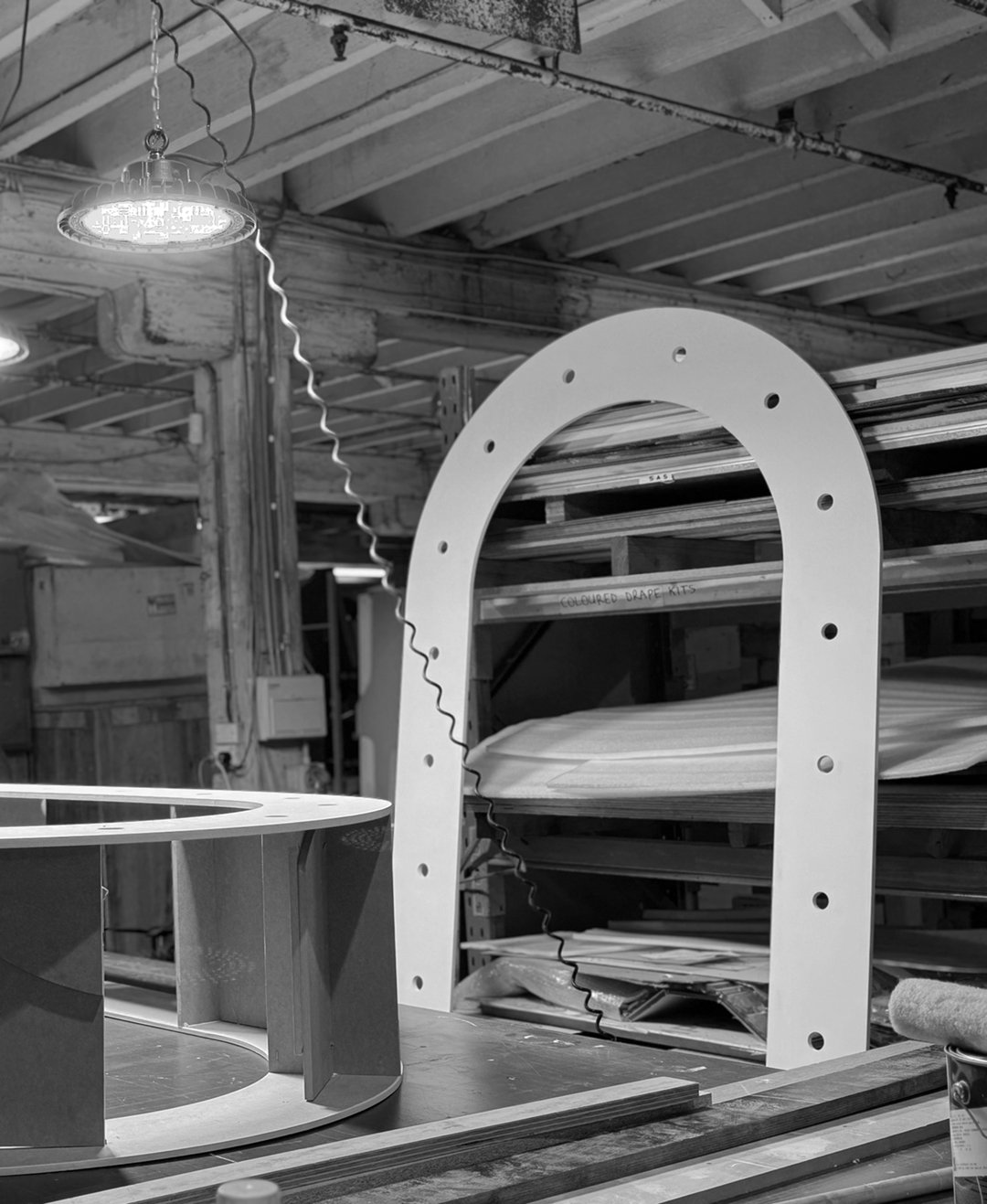 A woodworking workshop with a large white arch-shaped frame with bolt holes, a circular metal structure in the foreground, and shelves filled with wood planks and supplies in the background.