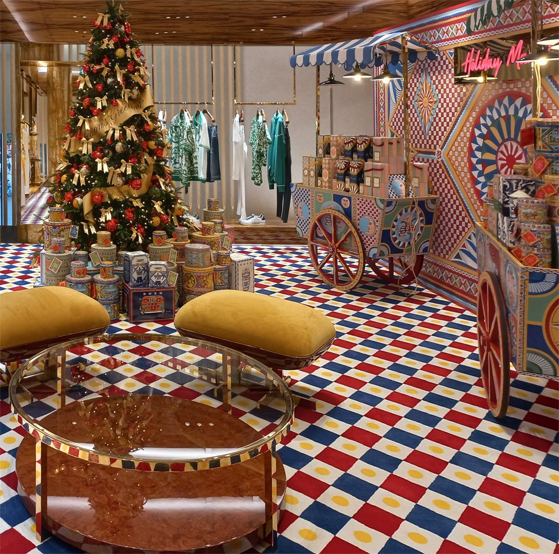 Custom high end luxury retail brands window dressing, installation.Decorated Christmas tree with gold ribbons, ornaments, patterned gift boxes in a colorful retail store with clothing racks, a vintage-style cart, geometric patterns. HR Productions