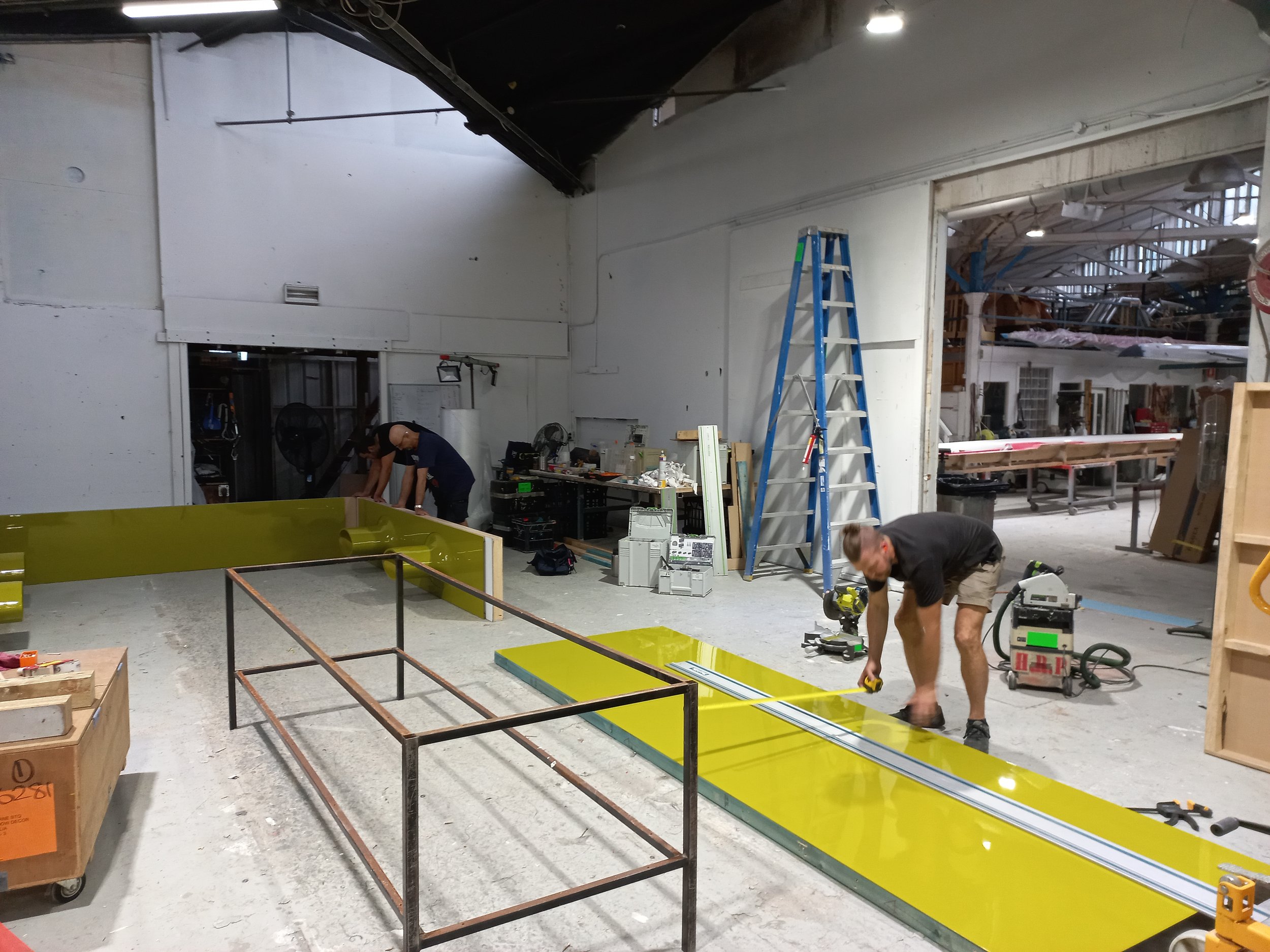 Workers assembling or repairing a large structure in a workshop with tools, a ladder, and various equipment around. HR Productions workshop space. Hire Custom built, props, sydney