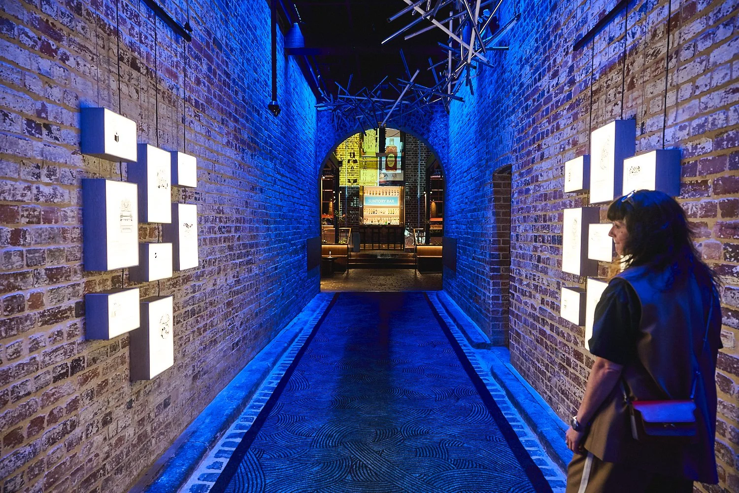 A narrow brick heritage space illuminated with blue lighting, featuring wall-mounted custom built light boxes, branded hospitality, Suntory luxury brand for CMMS. Sydney Production. HR Productions