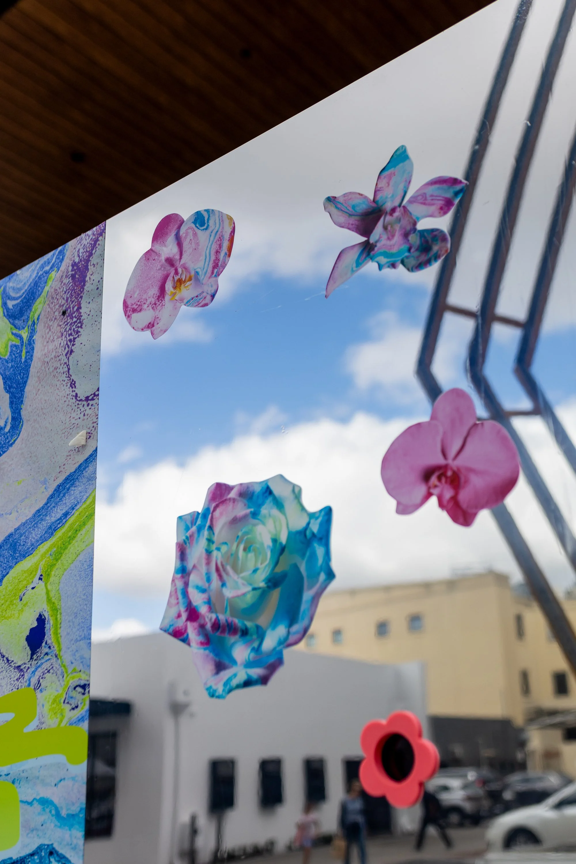Colorful paper flowers and artwork hanging on a wire outdoors with a cloudy sky and a building in the background. Acid FLWRS custom built outdoor installation. HRP Productions