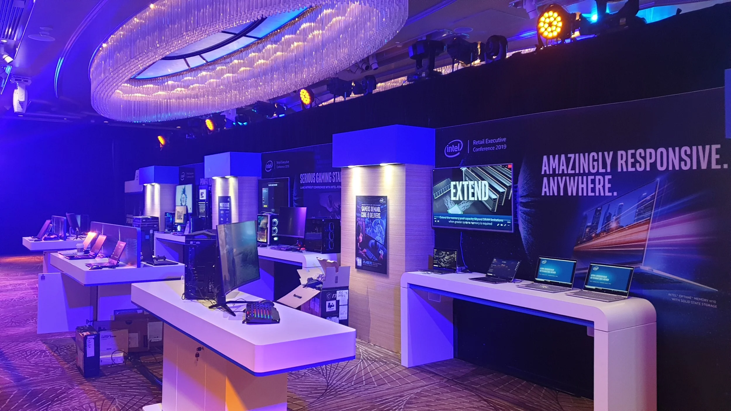 Exhibition Stand custom built. Technology display area with multiple computer monitors, gaming hardware, and promotional banners illuminated by blue and purple lighting. HR Productions