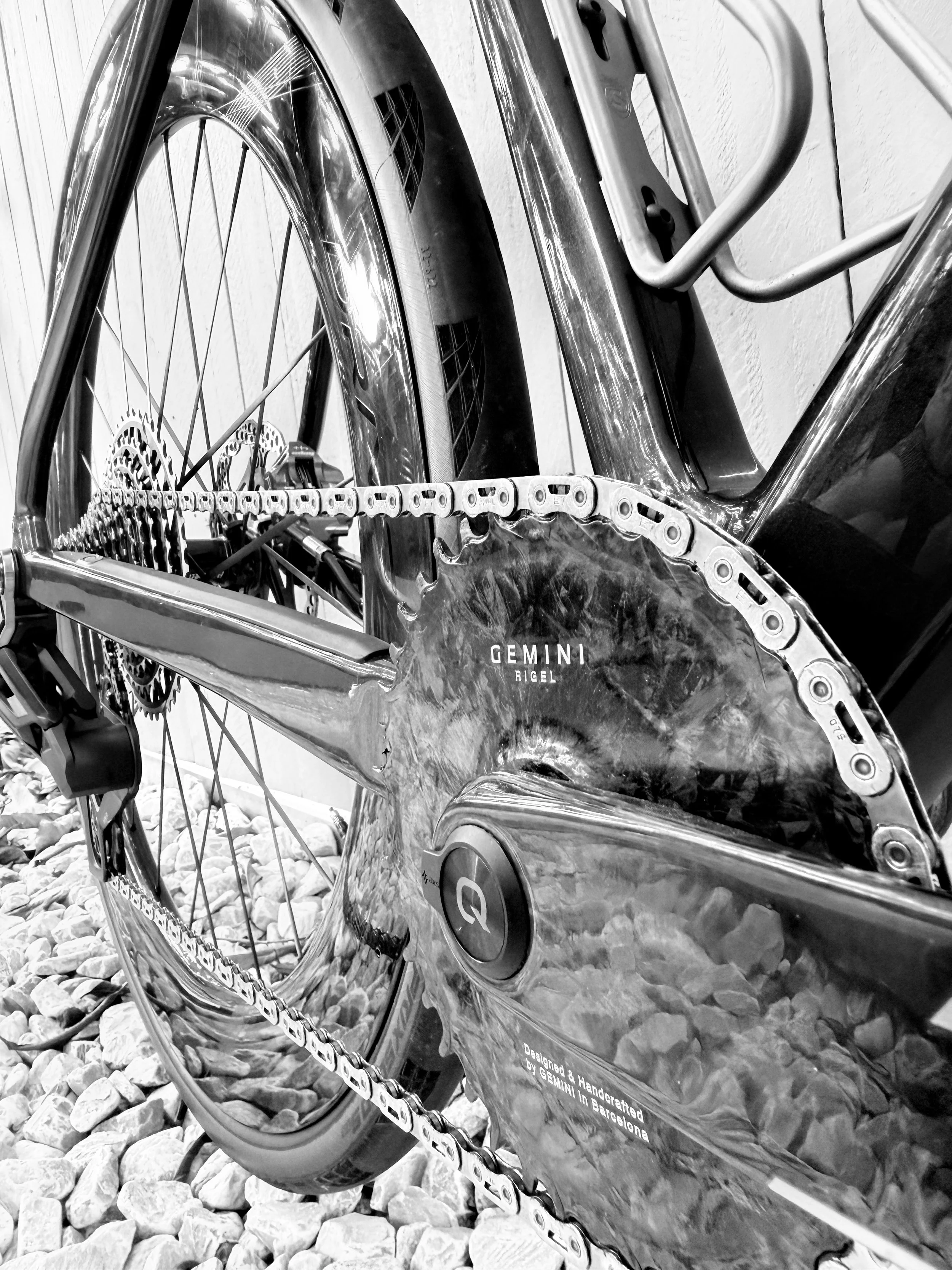 Close-up of a black and silver Gemini Rigel bicycle frame and wheel, with visible chain and gears, on a gravel ground.