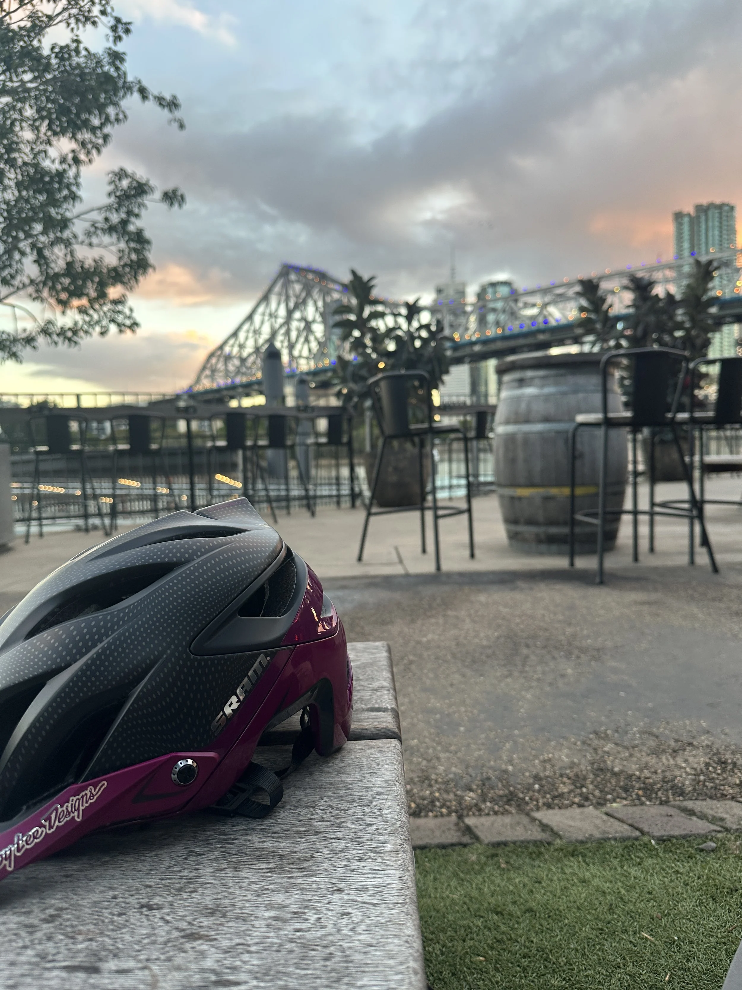 Close-up of a pink and black bicycle helmet sitting on a bench in an outdoor seating area. In the background, there are empty tables and chairs with a large barrel-style table, some trees, and a bridge with city buildings against a cloudy sky at dusk.