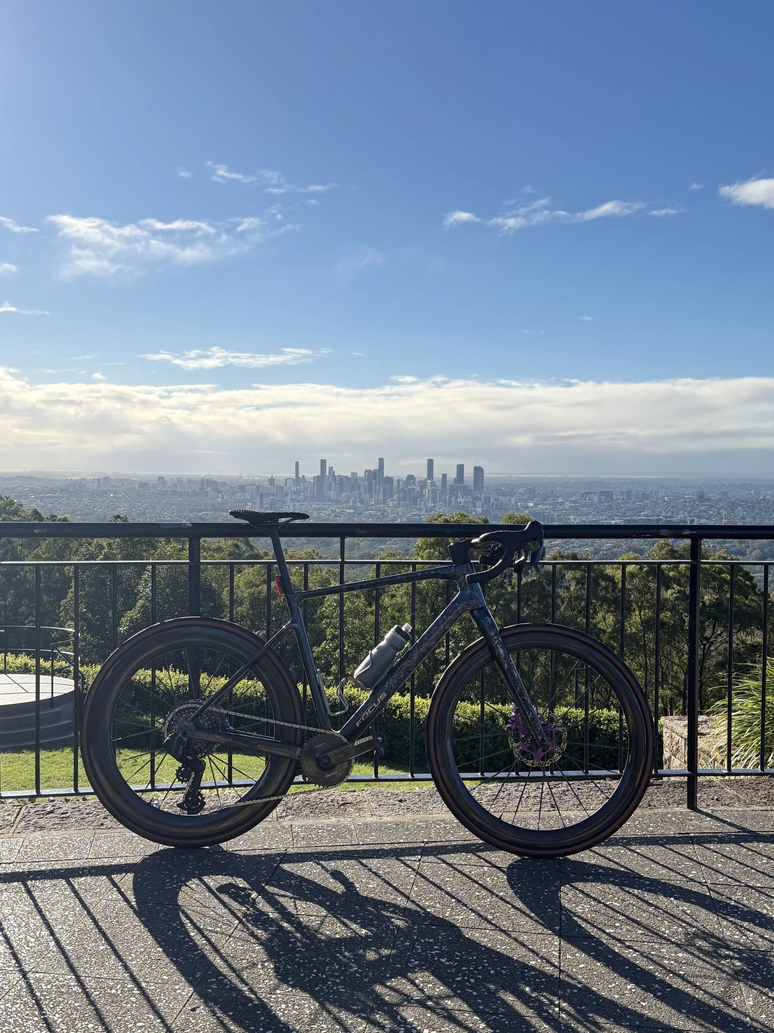 A bicycle parked against a black metal railing with a city skyline and blue sky with clouds in the background.