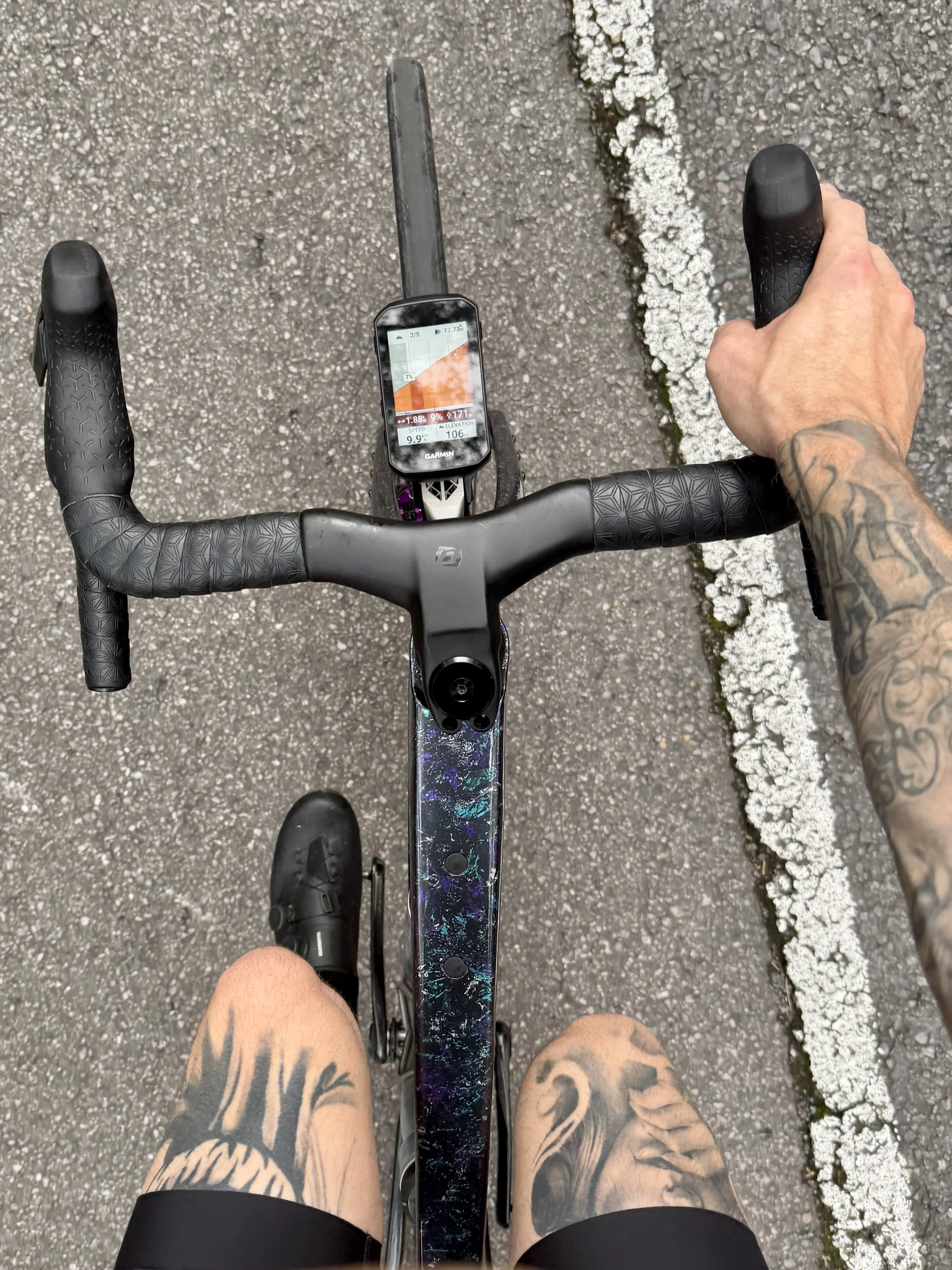 Overhead view of a person riding a bicycle on a paved road with a white line on the right edge. The person's arms and tattooed legs are visible, and a Garmin bike computer is mounted on the handlebar.