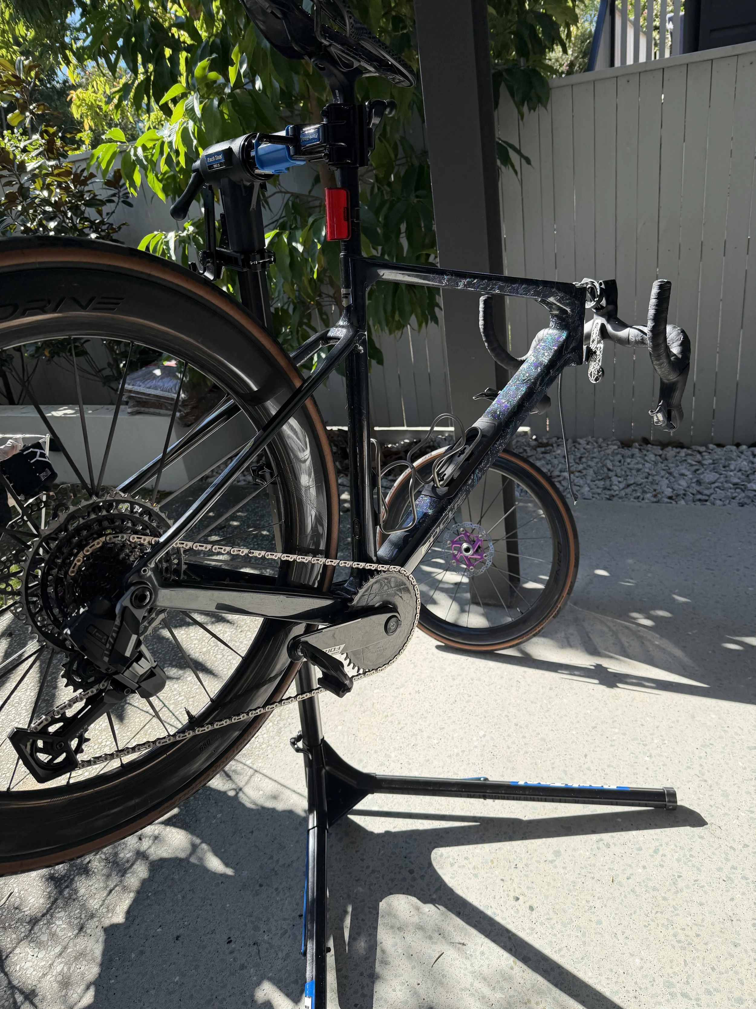 A road bike with a black frame and cork tires, placed on a bicycle stand outdoors with green trees and a white fence in the background.