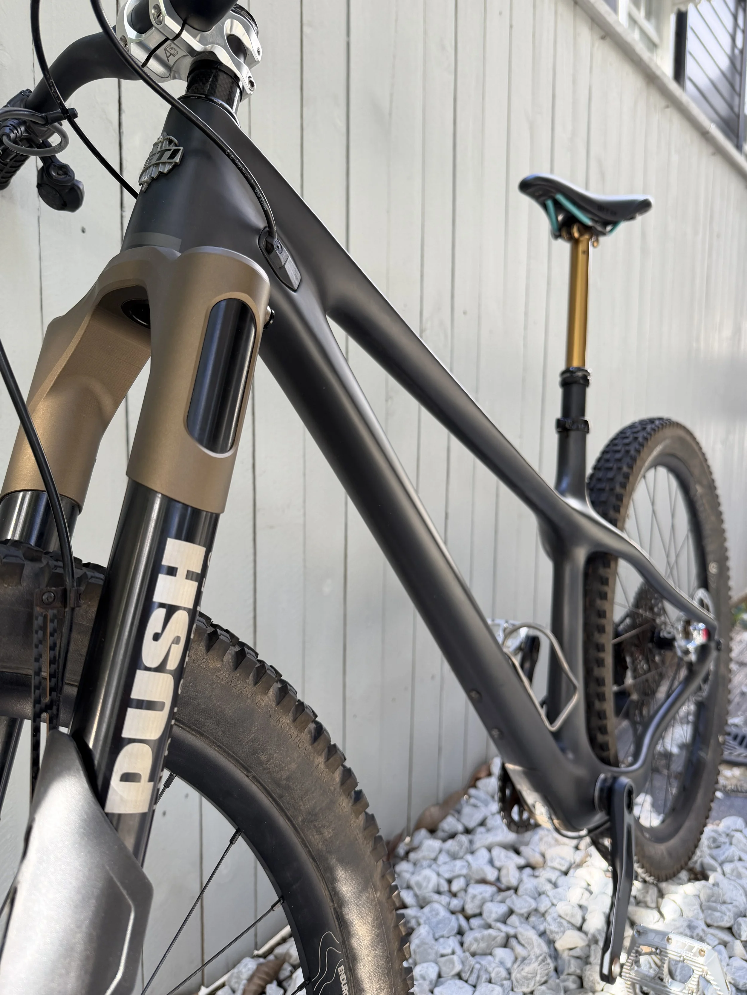 Close-up of a mountain bike leaning against a light-colored wooden fence, showing the front suspension fork, part of the frame, and the rear wheel with a knobby tire, set on a gravel surface.