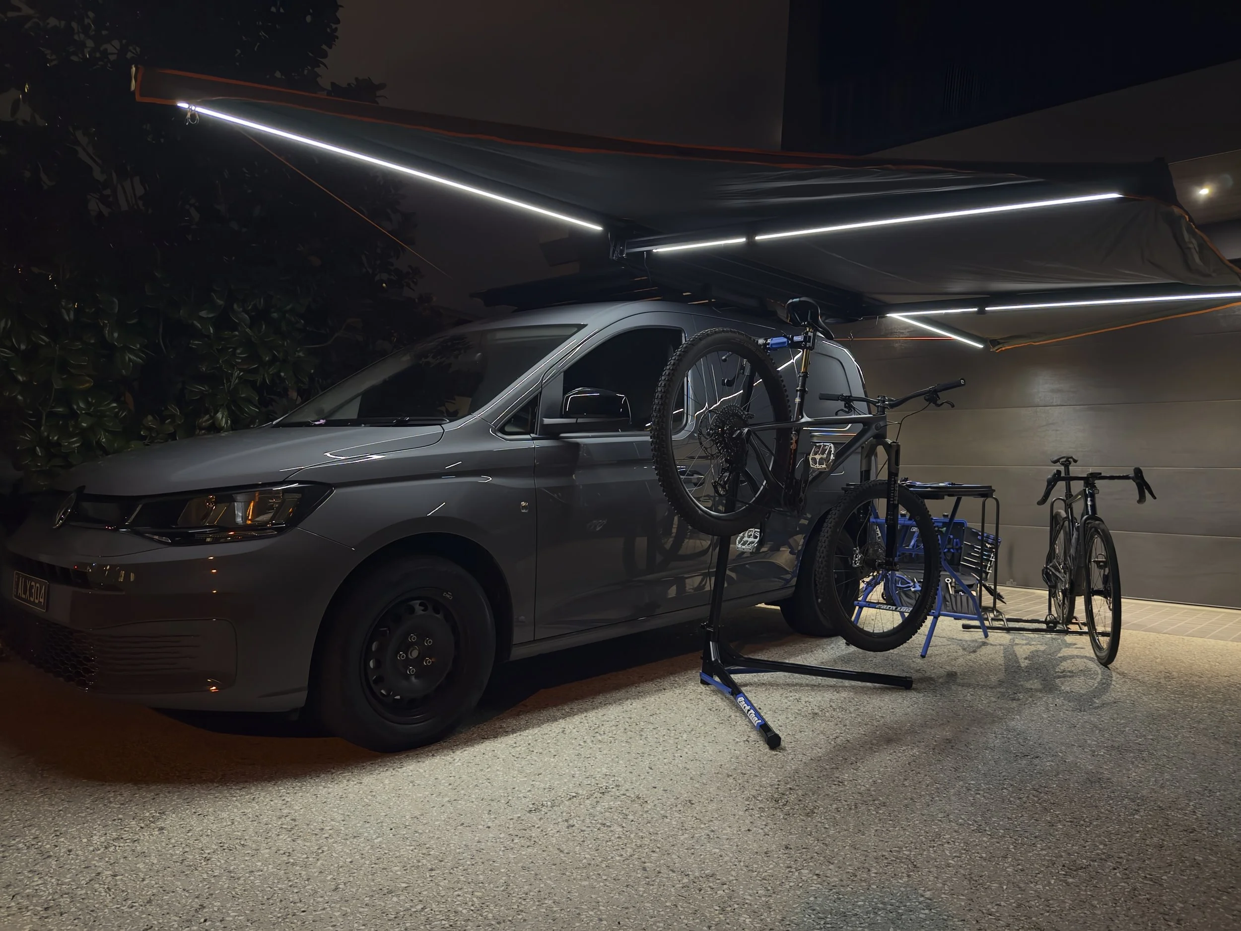Nighttime scene showing a silver car parked in a garage, with multiple bicycles stored on a rack above the car, illuminated by overhead lights.