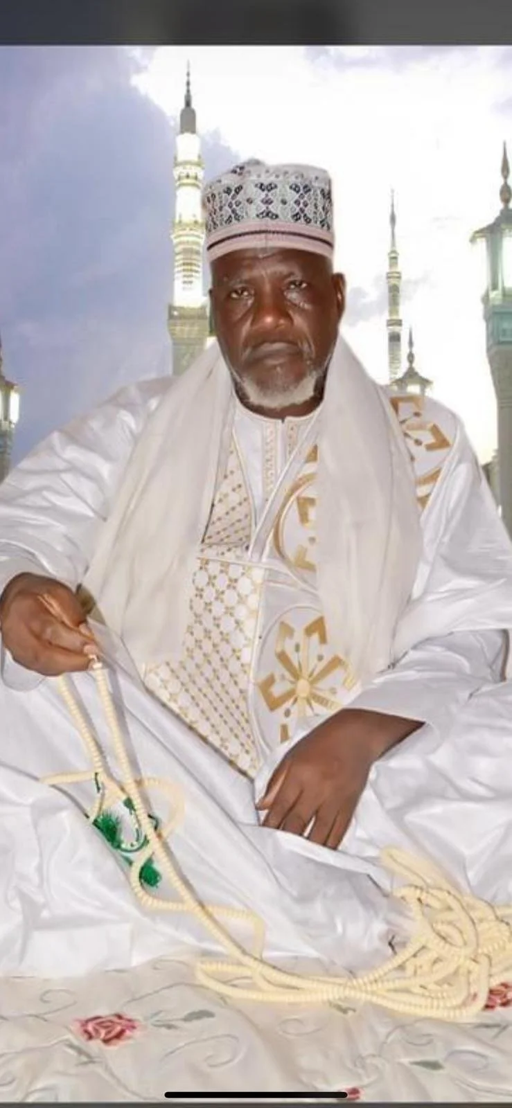 Homme âgé en vêtements blancs traditionnels, portant un calotte brodée, assis dans un décor de mosquées avec des minarets en arrière-plan.