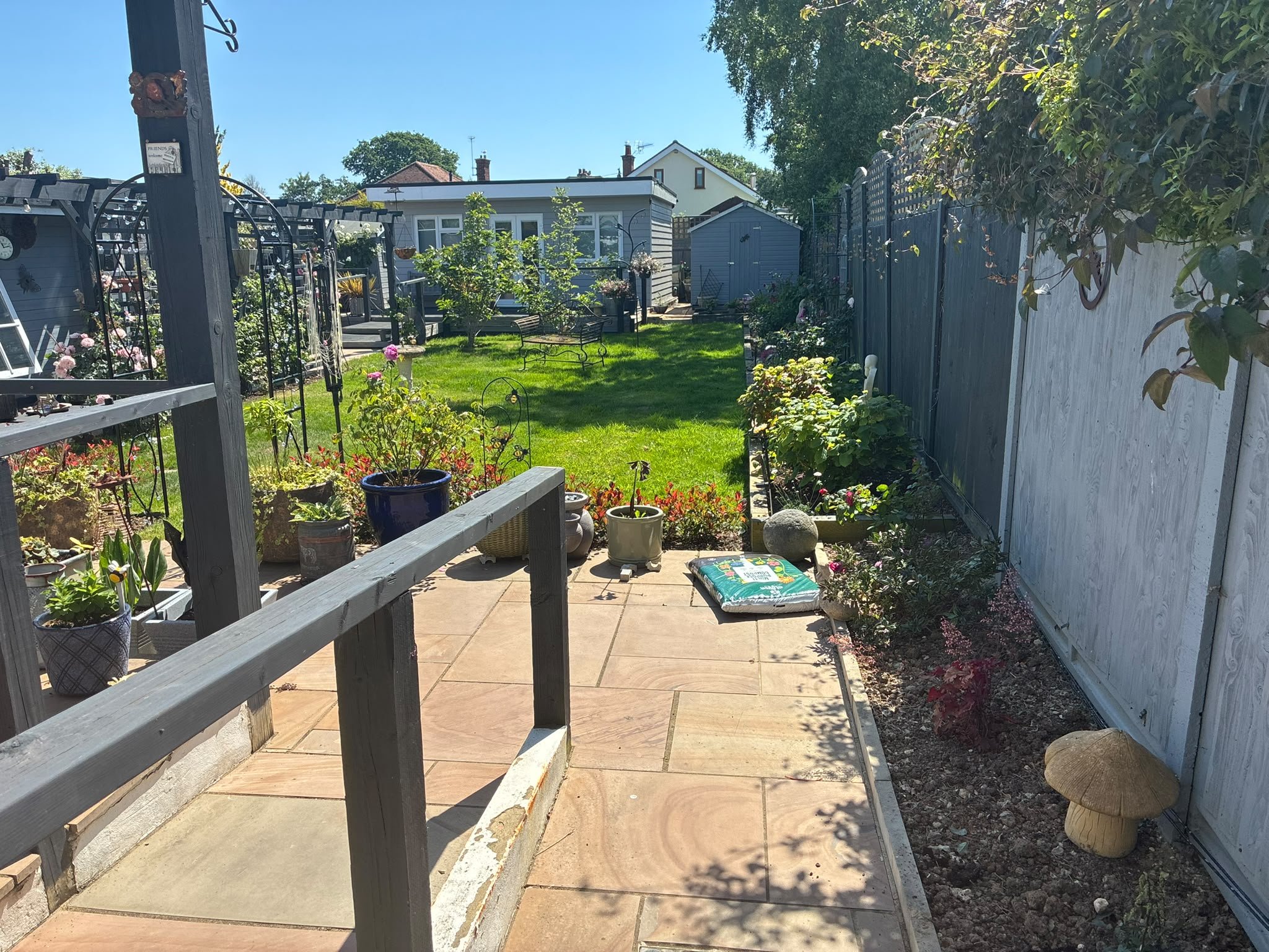 A backyard garden with a patio, a grassy area, trees, and a shed. Decorative plants and flowers are in pots and beds, with a bench and garden tools visible.