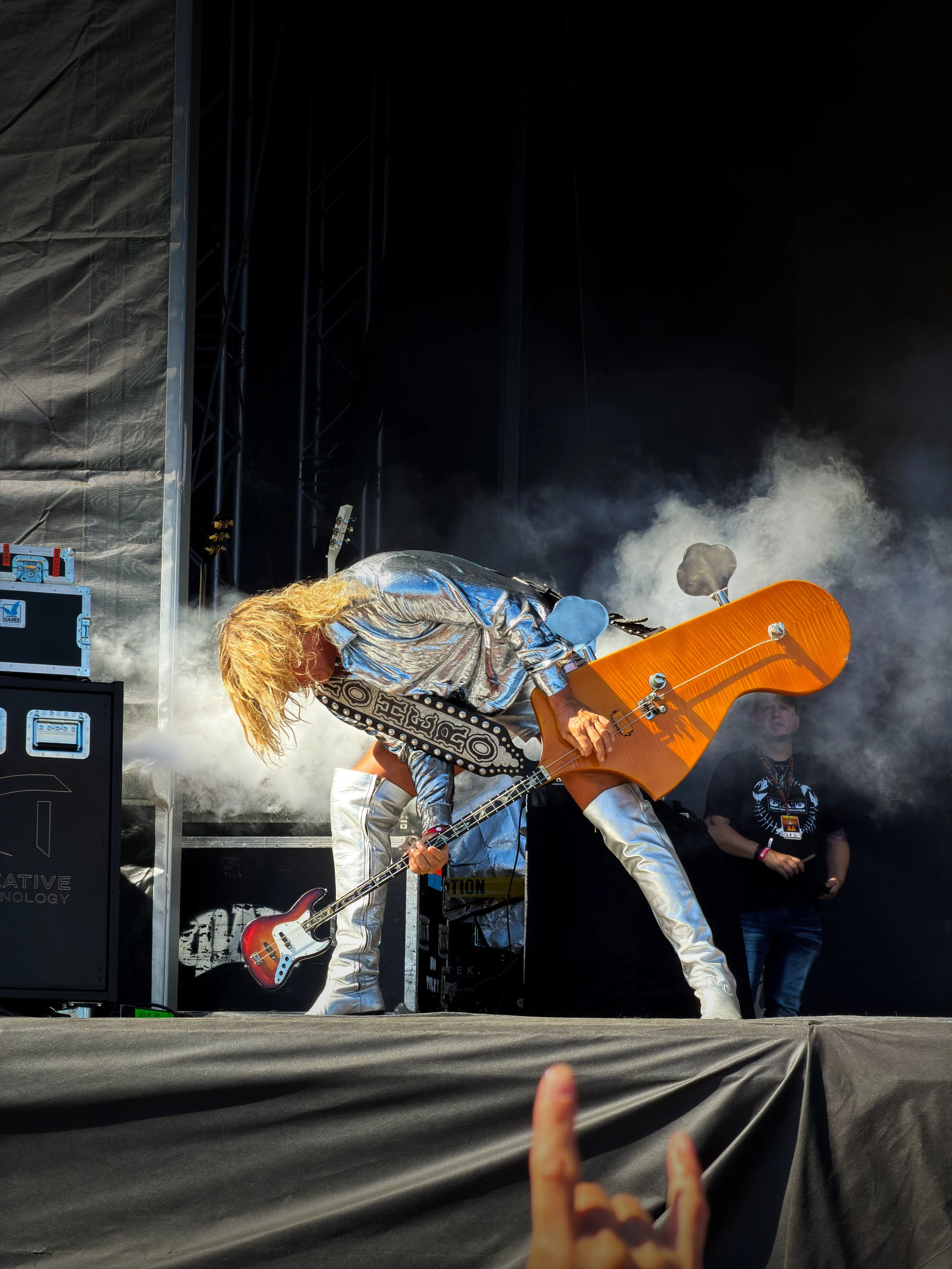 A live music performance on stage featuring a guitarist in a shiny silver outfit, playing an orange electric guitar. Smoke effects and stage equipment are visible in the background, with an audience member’s hand making a rock and roll gesture in the foreground.