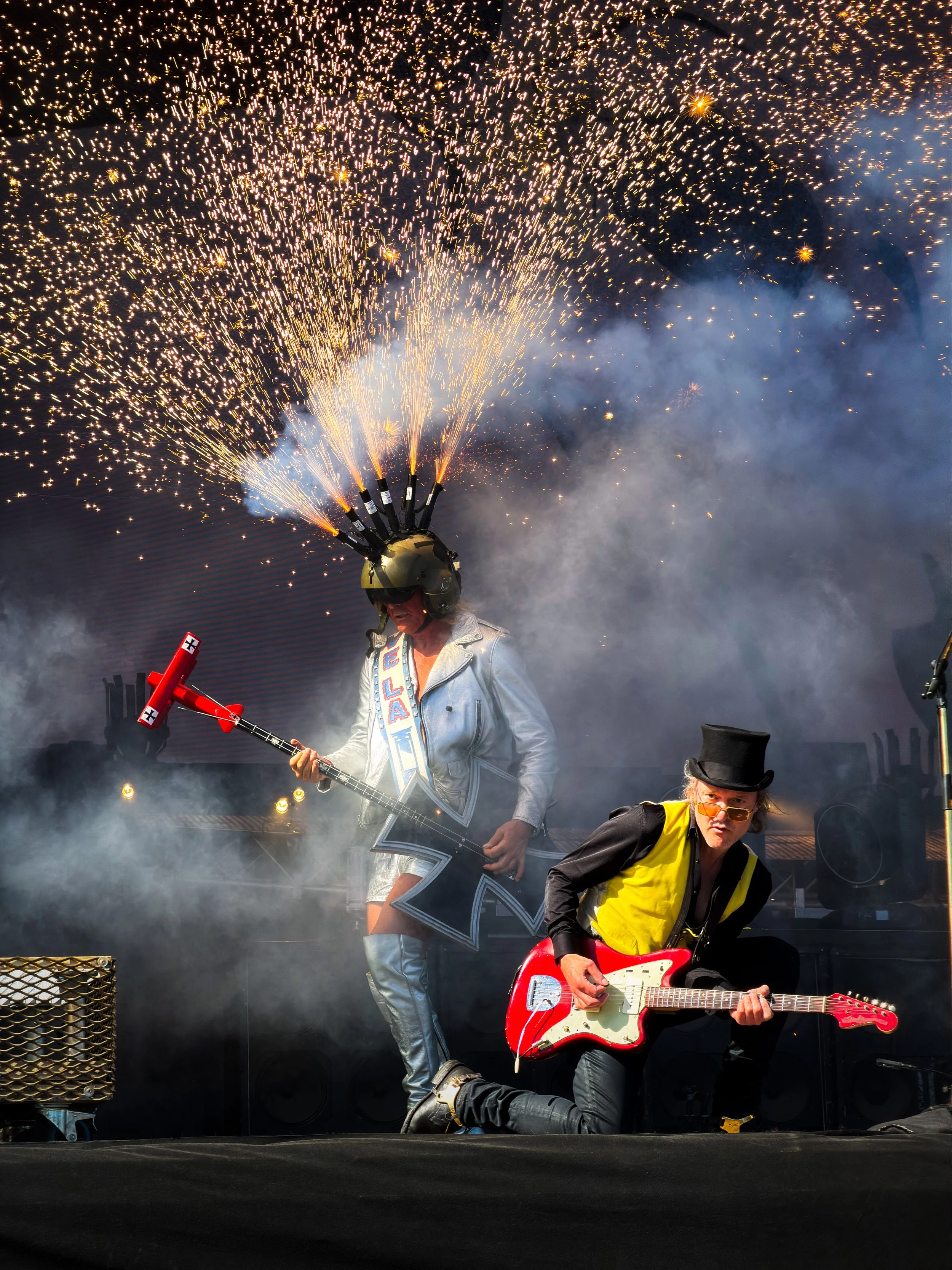 Musicians performing on stage during a fireworks display, with sparks flying from a handheld rocket launcher and another playing an electric guitar, a woman in a leather jacket and yellow vest in the foreground.
