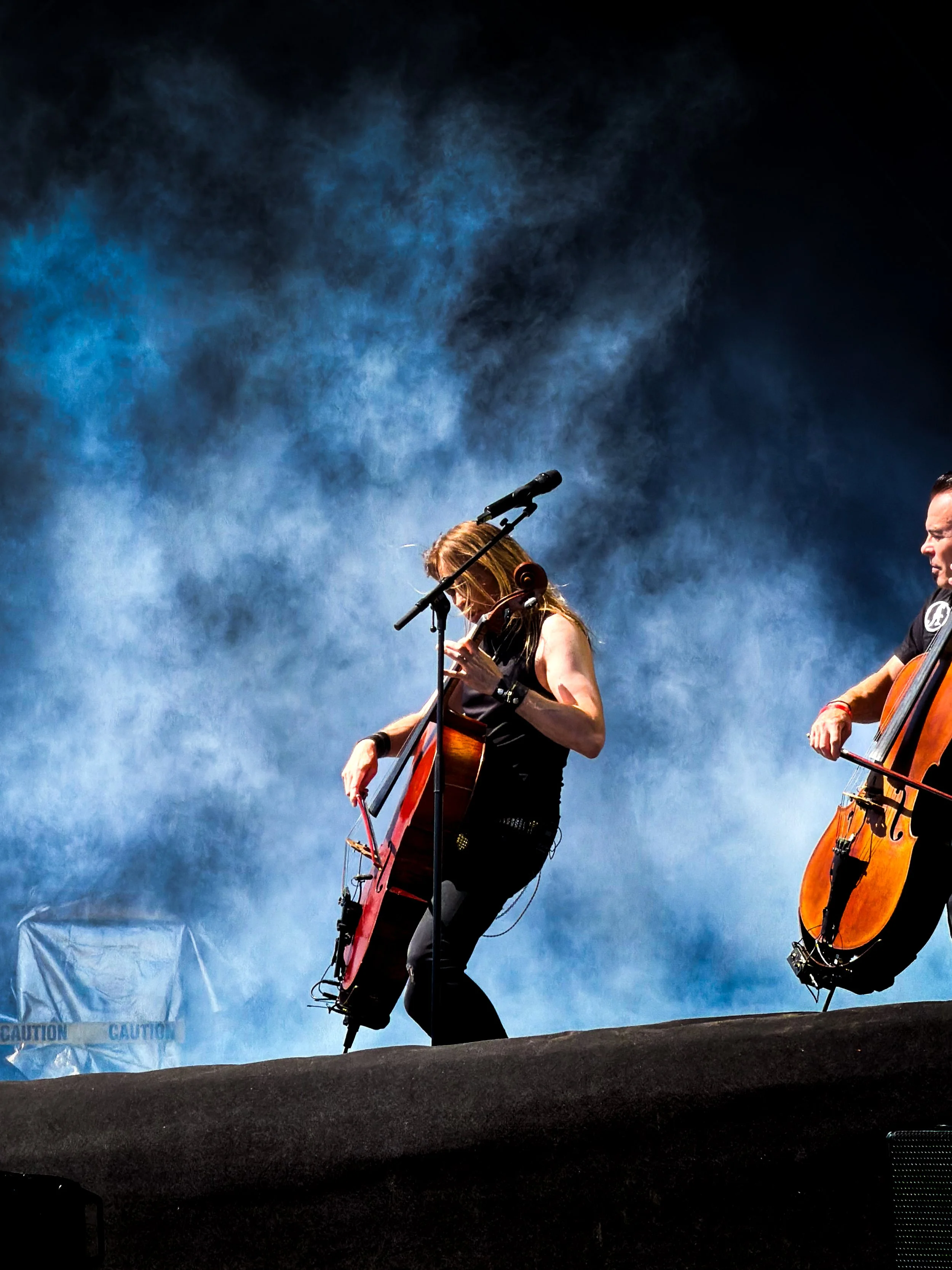 Two musicians playing guitars on stage with blue smoky background, one woman with long hair, sleeveless top, and another man with short hair, partly visible.
