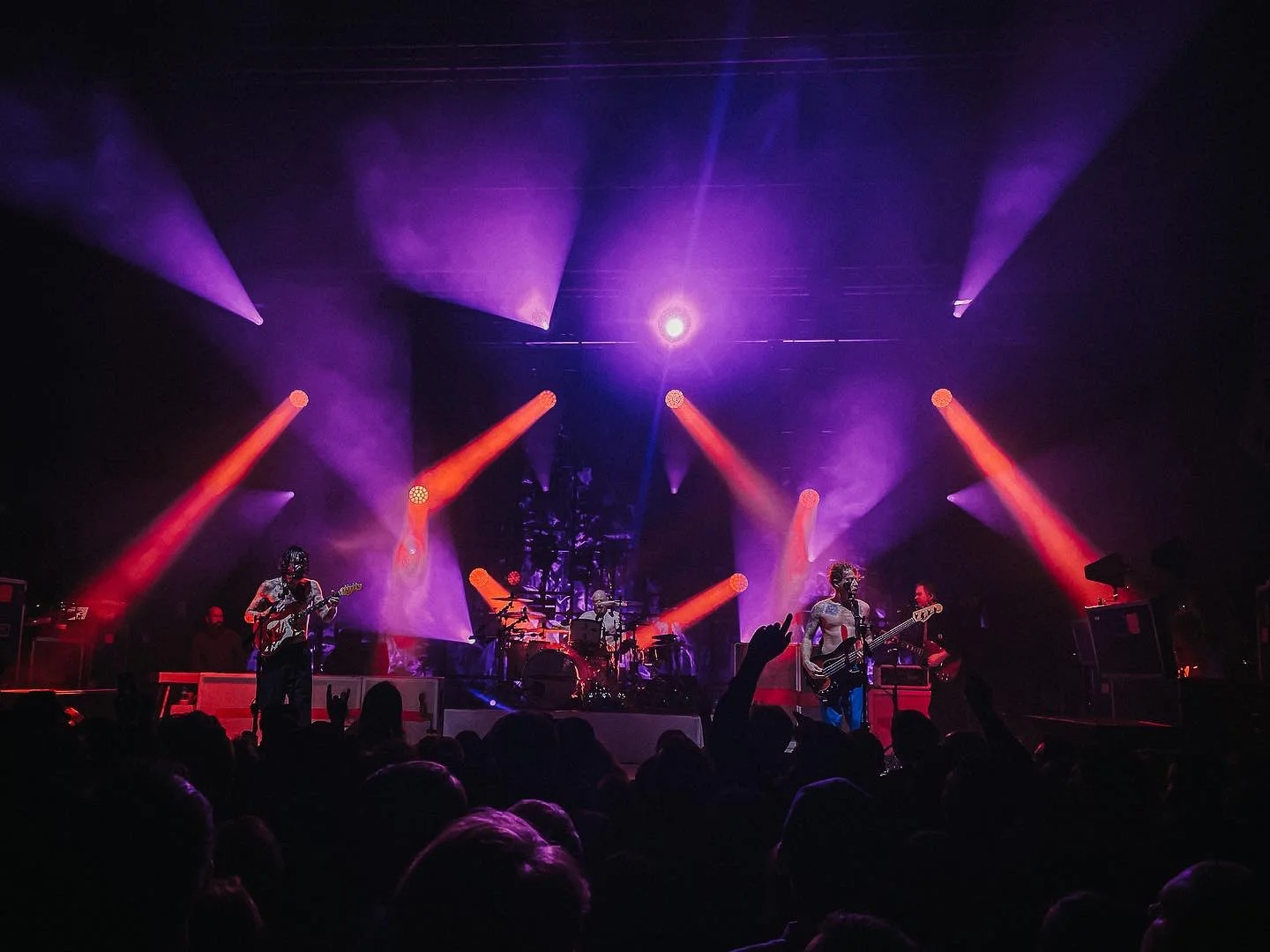 A live music concert with a band performing on stage under purple and orange stage lights, with an audience in the foreground.
