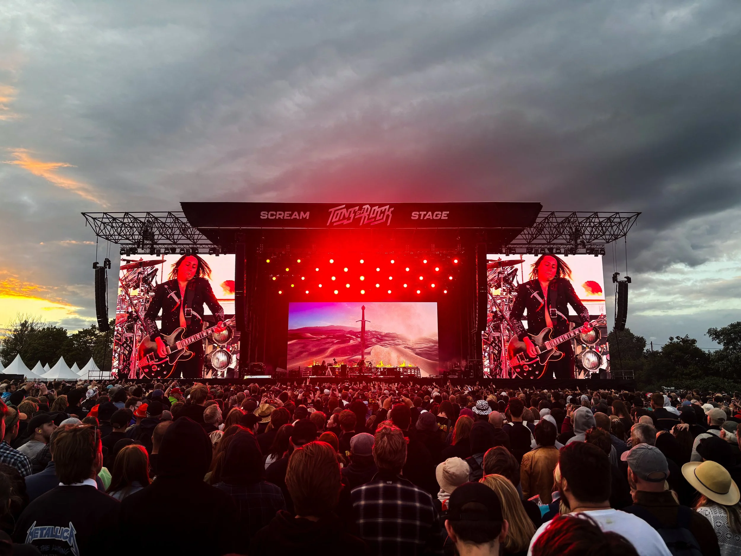 A large crowd of people attending an outdoor concert at dusk, watching a stage with a performer playing a guitar and a band, with a cloudy sky overhead.