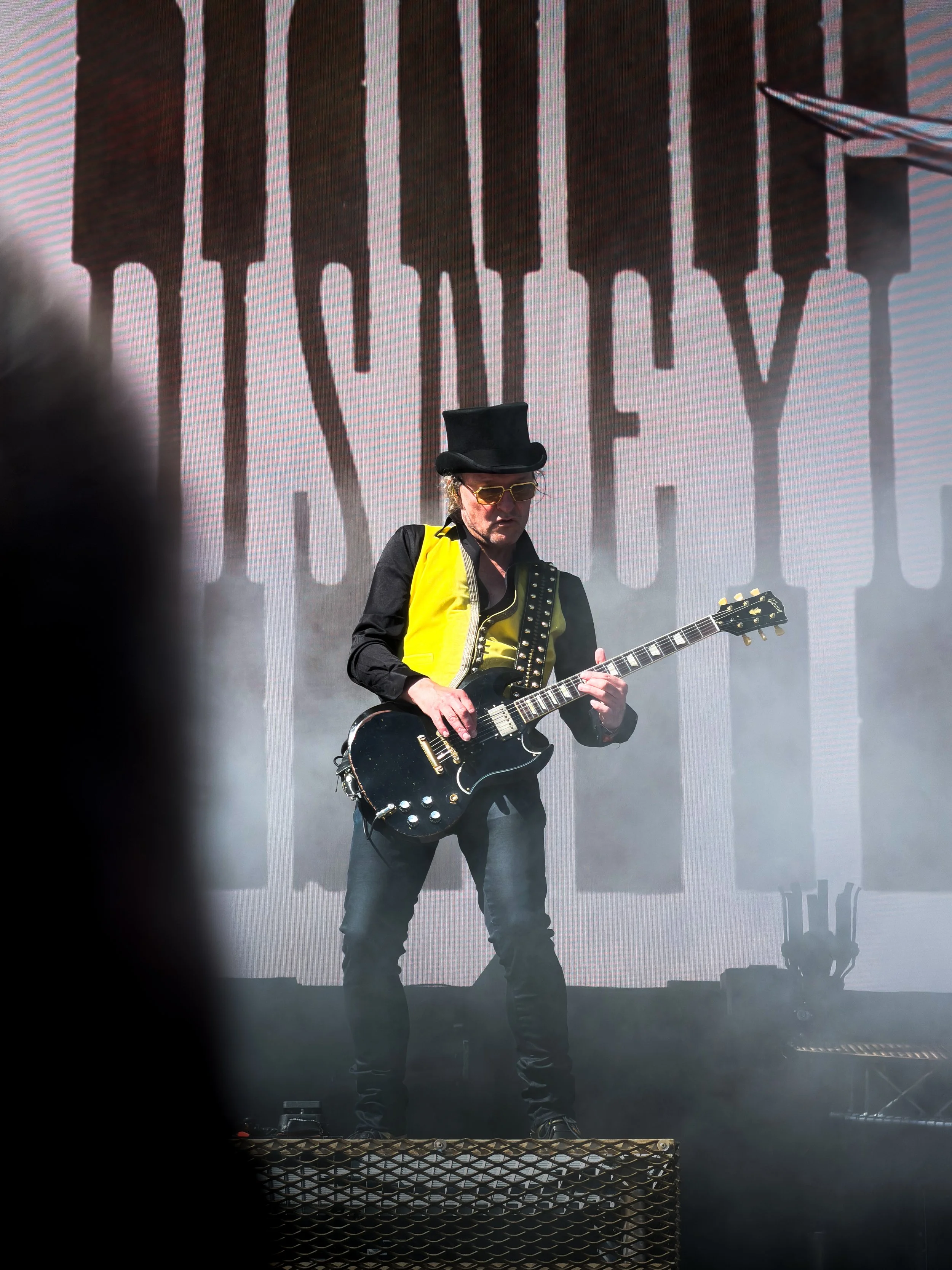 A person wearing a black top hat, sunglasses, and a yellow and black jacket playing an electric guitar on stage, with a large screen behind displaying partially visible text.