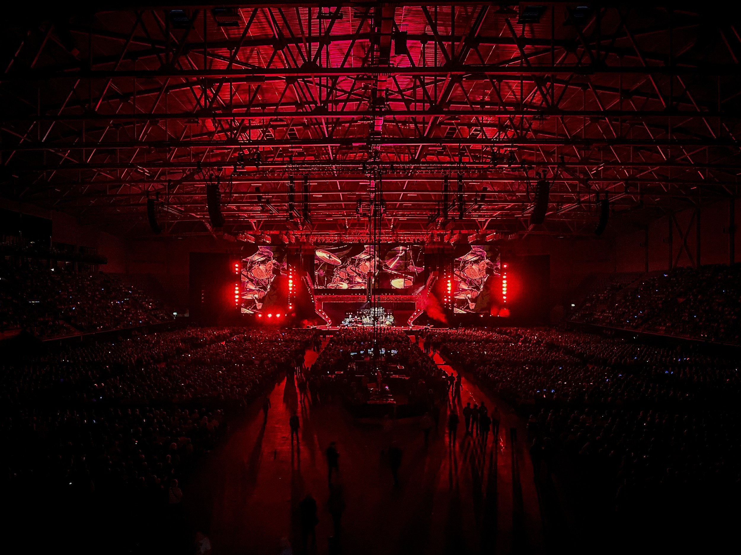 Concert stage illuminated with red lights, large screens displaying abstract images, and a crowd of attendees in the dark.