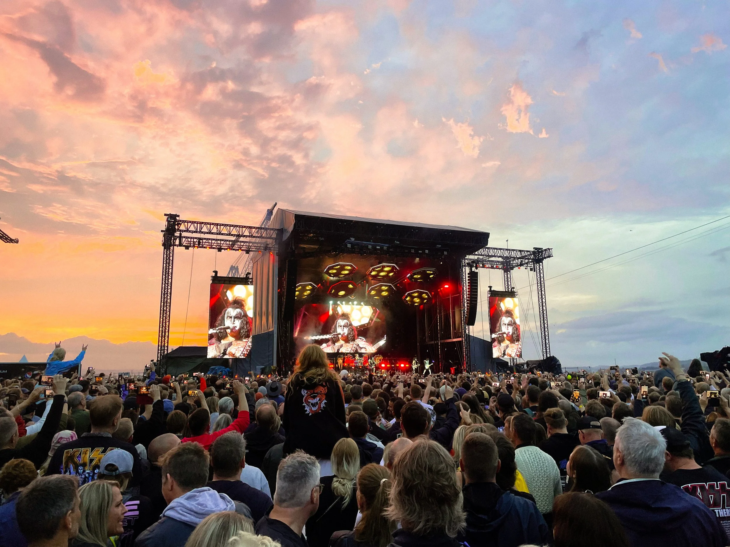 A large crowd of people gathered outdoors at a concert or festival during sunset. The stage with bright lights and screens displaying performers is in the background. The sky is partly cloudy with shades of pink and orange.