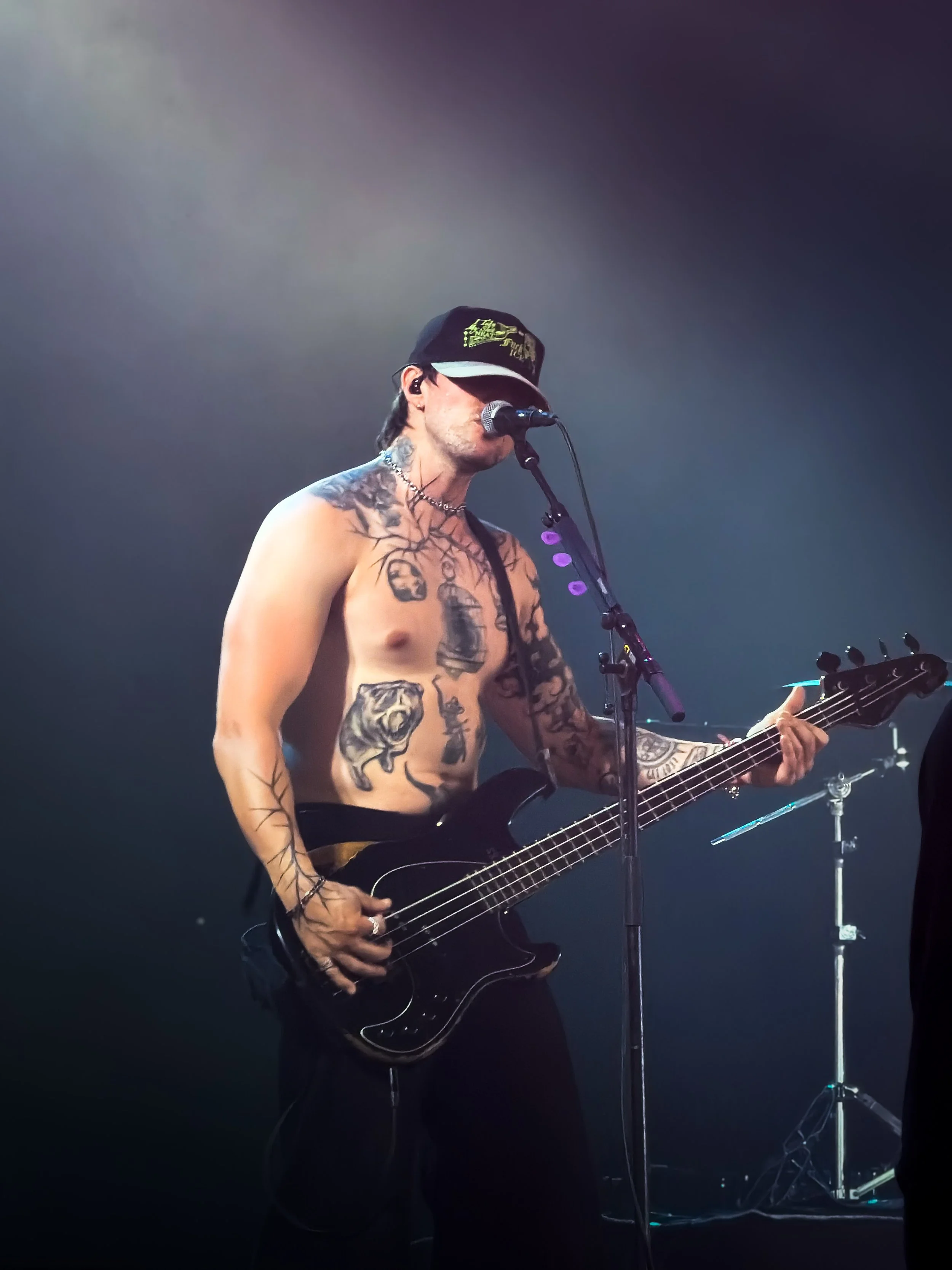A shirtless male musician with tattoos, wearing a black baseball cap, singing into a microphone while playing a black electric bass guitar on stage with dark lighting.
