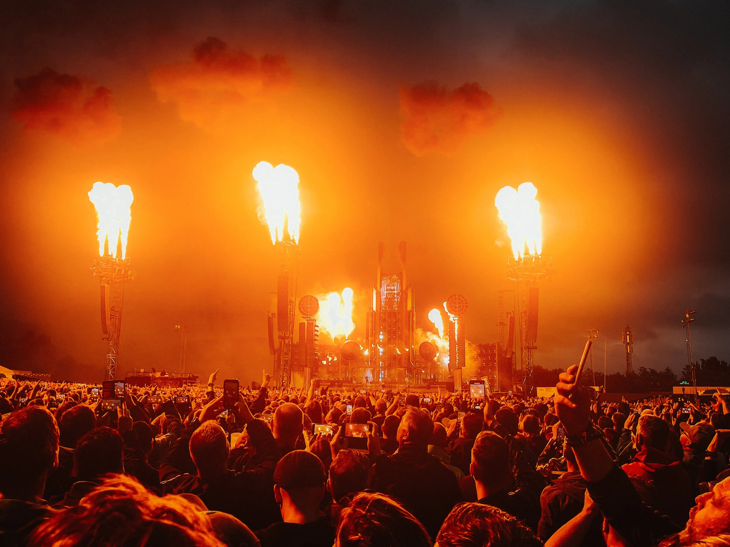 A large crowd attending a concert or event at night with a stage in the background. The stage is illuminated with bright flames shooting into the air, and behind it, there are large speakers and lighting equipment. Many people in the crowd are capturing the moment on their phones.